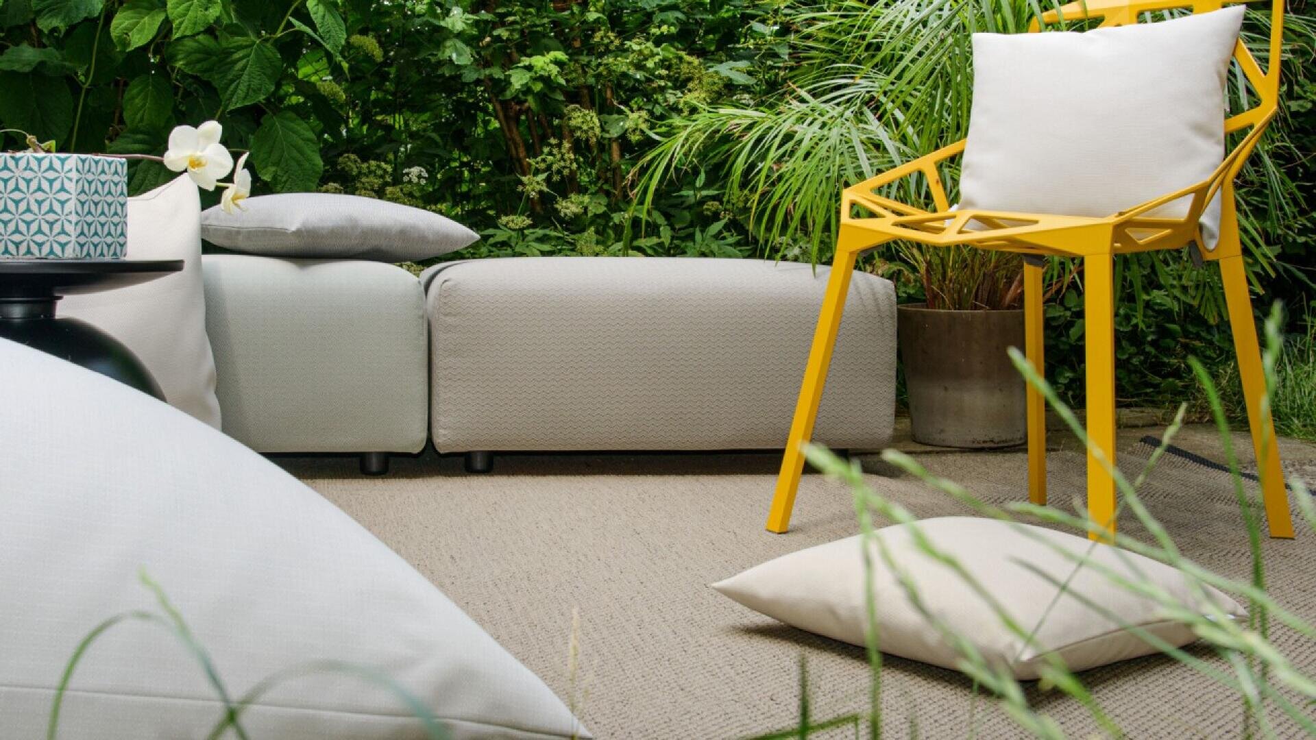 A modern outdoor seating area with light gray cushions, a yellow geometric chair, and white pillows, surrounded by lush green plants on a neutral rug.