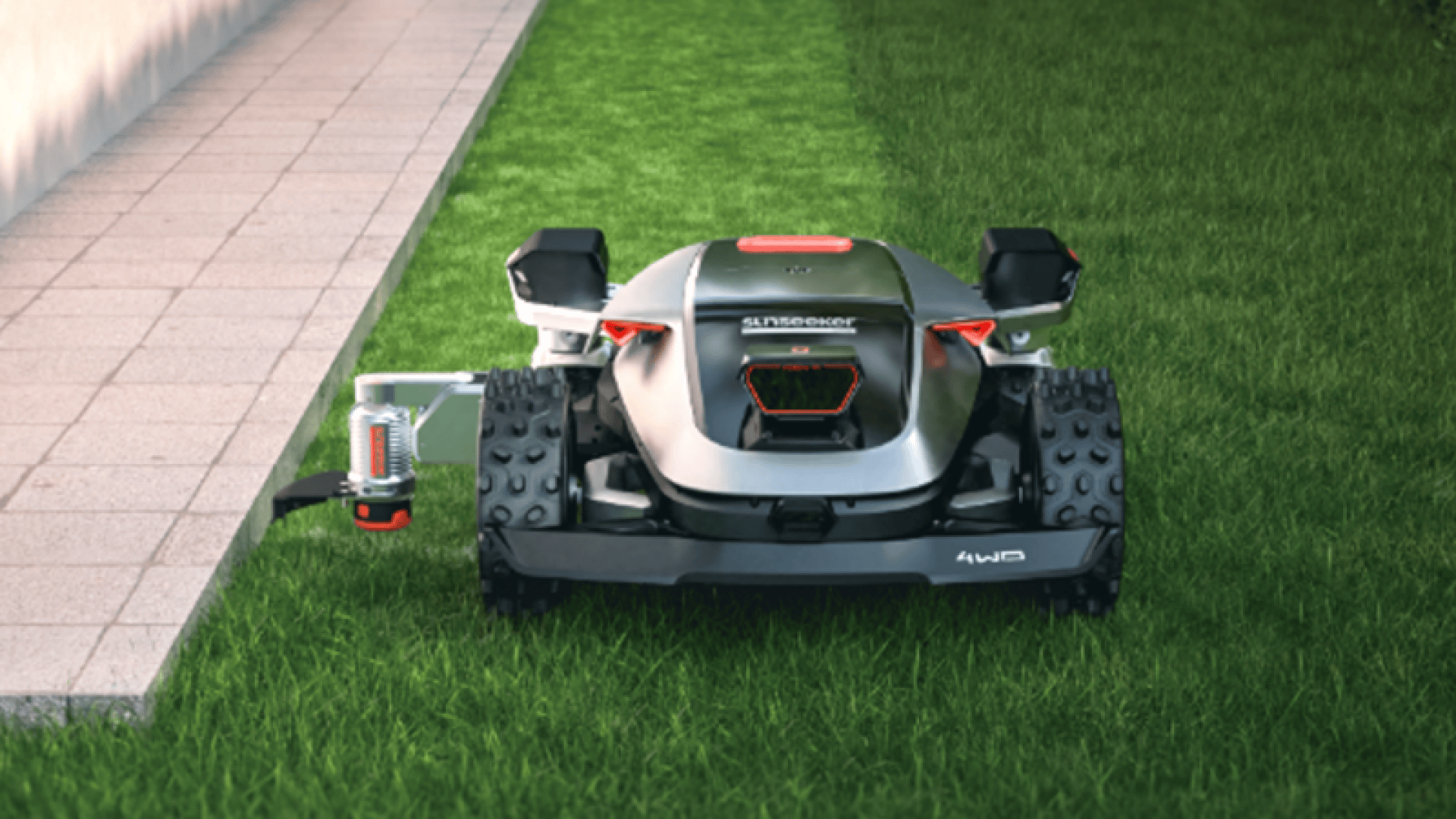 A robotic lawn mower with robust wheels and a metal housing mows grass next to a paved sidewalk. The mower is equipped with four-wheel drive and a visible sensor or antenna on the left-hand side.