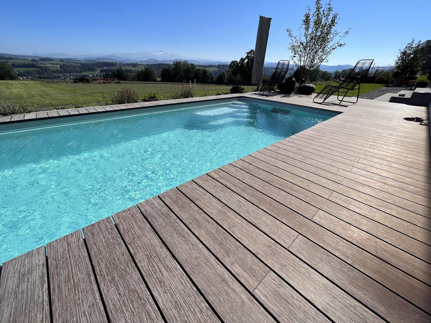 Ein klarer, blauer Swimmingpool mit Holzdeck, Liegestühlen und Sonnenschirm befindet sich im Freien, mit Blick auf ein grasbewachsenes Feld und entfernte Hügel unter einem hellen, sonnigen Himmel.