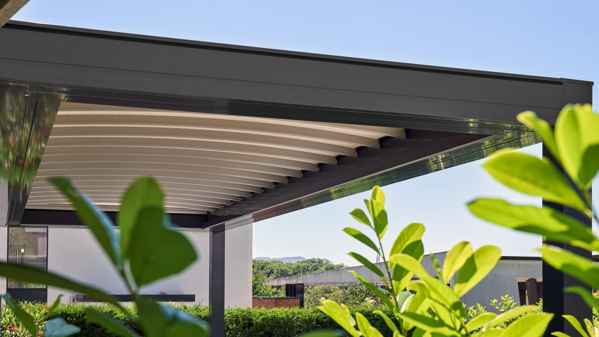 Eine moderne Pergola mit einem schlanken, dunklen Metallrahmen und einem weißen Lamellendach ist an einem sonnigen Tag abgebildet, umgeben von grünen Pflanzen und Blättern, mit blauem Himmel und Gebäuden im Hintergrund.
