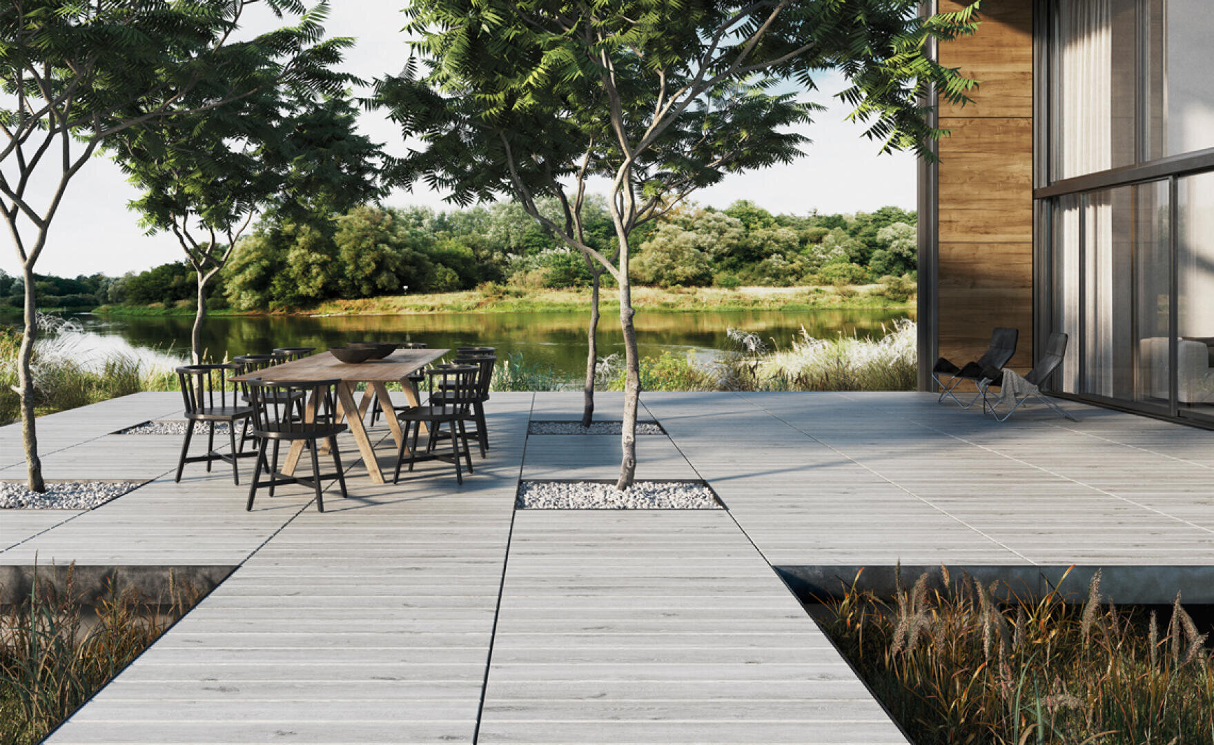 Moderne Holzterrasse mit schwarzen Stühlen und einem Tisch unter belaubten Bäumen, neben einem Gebäude mit Glaswänden, mit Blick auf einen ruhigen See und üppiges Grün im Hintergrund.