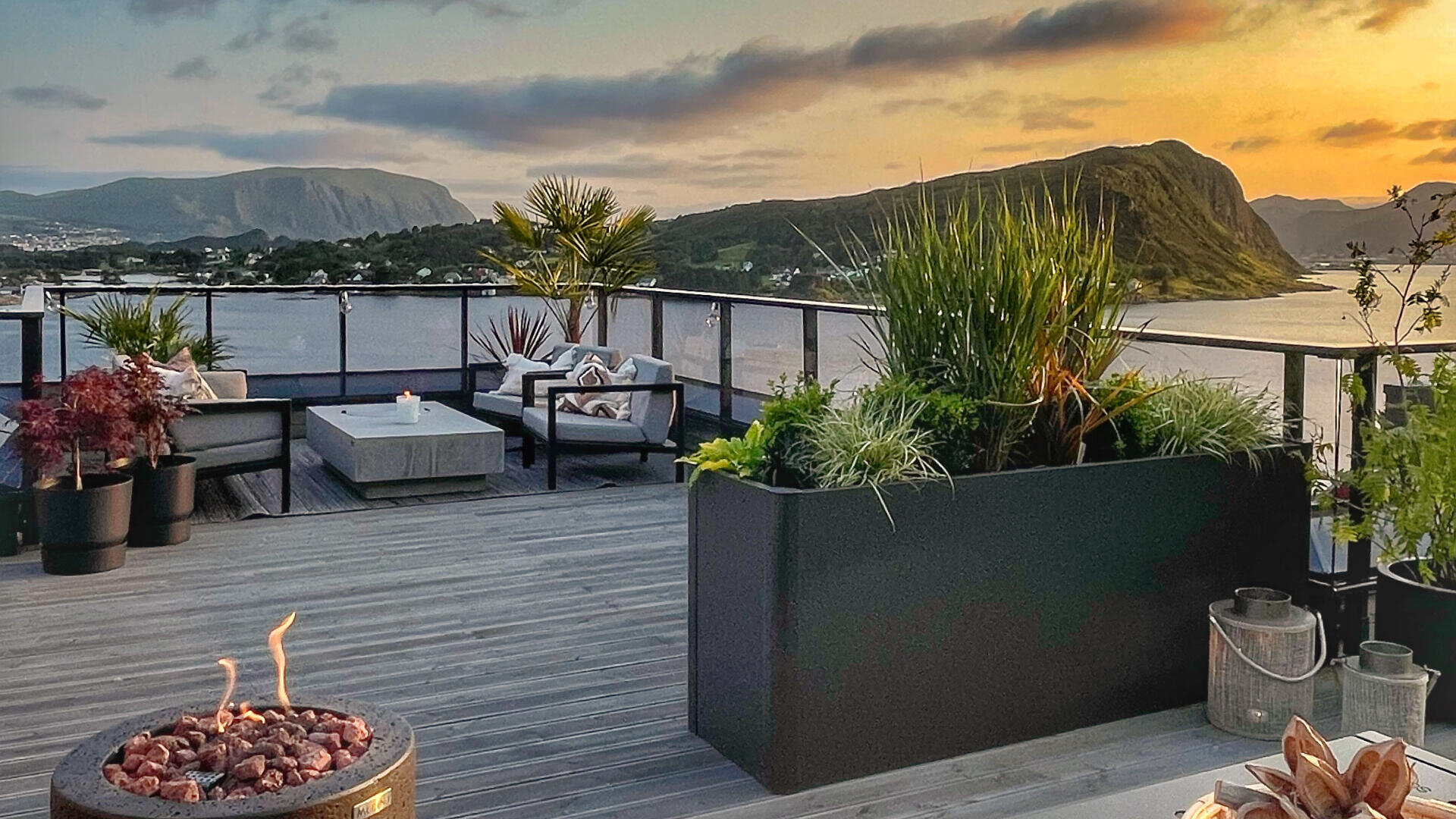 Dachterrasse mit modernen Terrassenmöbeln, Pflanzgefäßen und einer Feuerstelle mit Blick auf ein malerisches Gewässer und Berge bei Sonnenuntergang. Der Himmel ist teilweise bewölkt mit warmem, goldenem Licht.