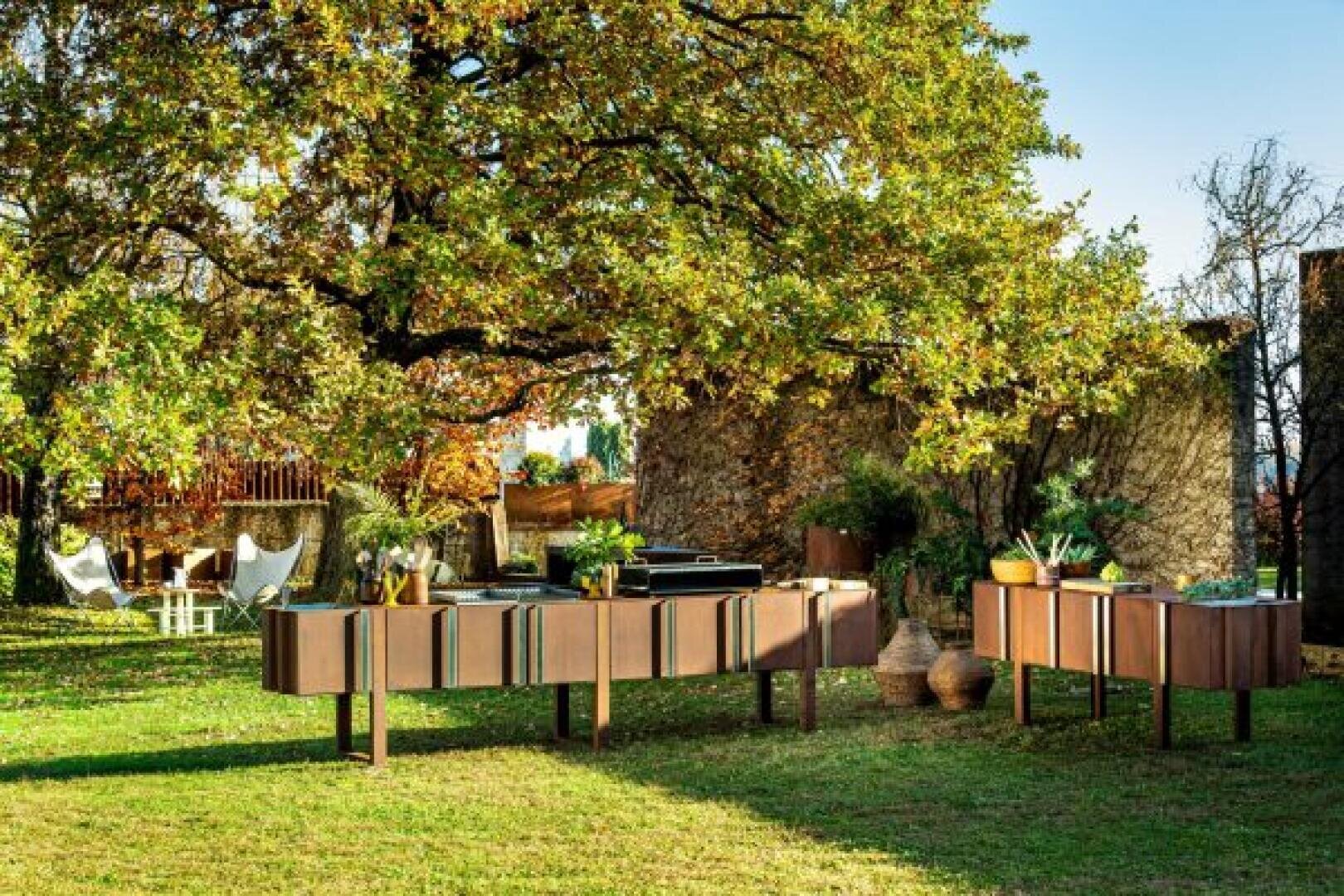 Outdoor kitchen setup with modern wooden cabinets and countertops beneath a large tree, surrounded by greenery, potted plants, and rustic stone walls; white outdoor chairs are visible in the background.