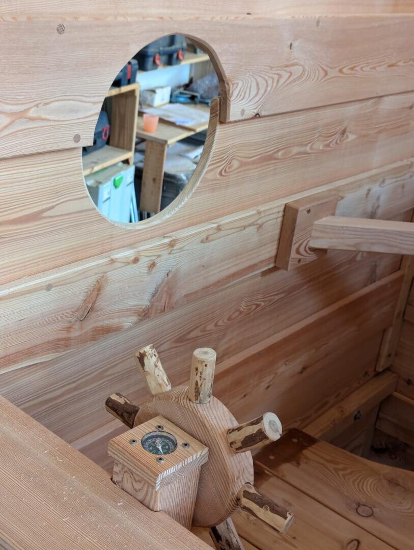 A close-up of a wooden ship’s wheel and compass inside a handmade wooden boat, with a round window showing a workshop in the background.
