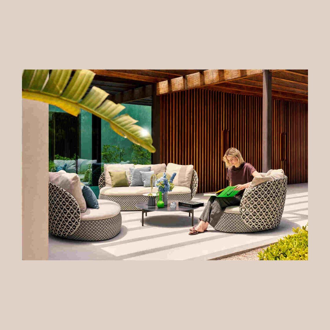 Eine Frau sitzt lesend auf einem gemusterten Gartenstuhl neben einem niedrigen Tisch mit Vasen, umgeben von passenden Sofas und üppigen Pflanzen auf einer modernen Terrasse mit Holzlatten und großen Fenstern.
