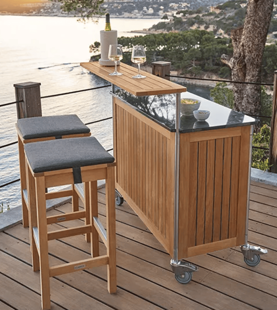 A wooden bar cart with a glass top and two wooden bar stools sits on a deck overlooking a coastal view at sunset. Wine glasses and a wine bottle are on the bar, and trees and water are visible in the background.