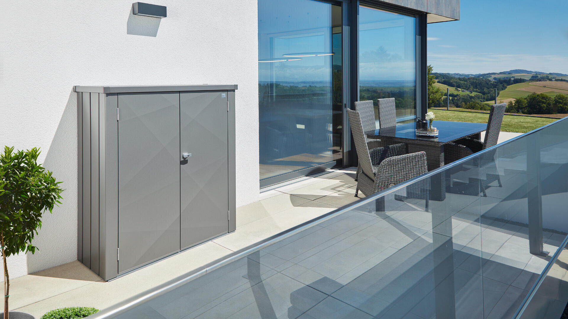 Moderne Außenterrasse mit einem grauen Metallschrank, einem Glasgeländer, einer Essgruppe aus Weidengeflecht, einer Topfpflanze und einer malerischen Aussicht auf die Landschaft, die sich in großen Glasfenstern spiegelt.