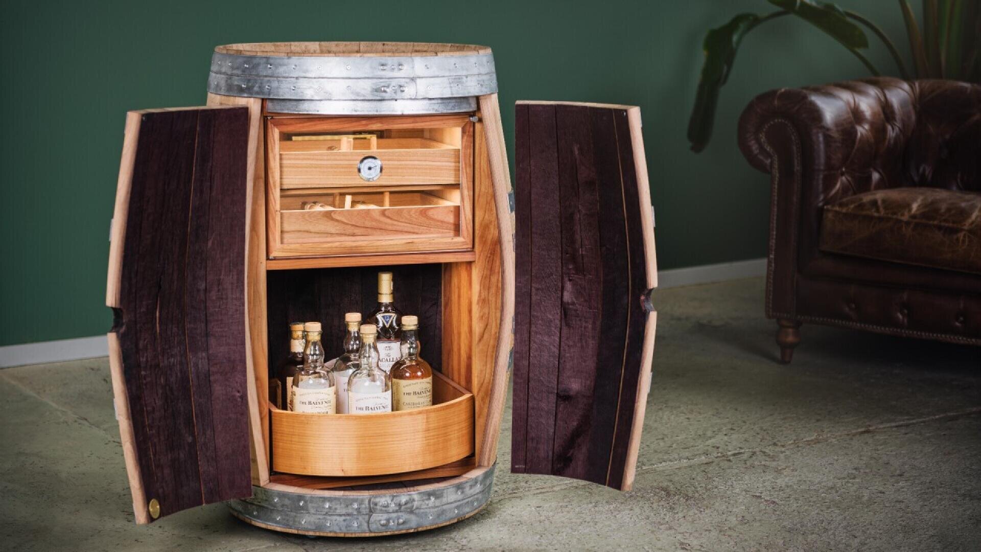 A wooden barrel cabinet with open doors reveals shelves holding bottles of liquor and a humidor drawer, standing on a concrete floor next to a brown leather sofa and a green wall with a plant.