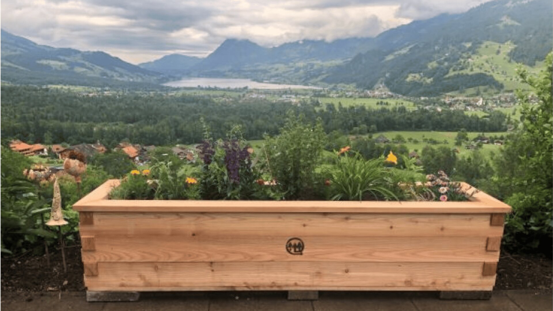 A wooden planter box filled with various flowers and plants sits on a patio, overlooking a lush green valley with mountains and a cloudy sky in the background.