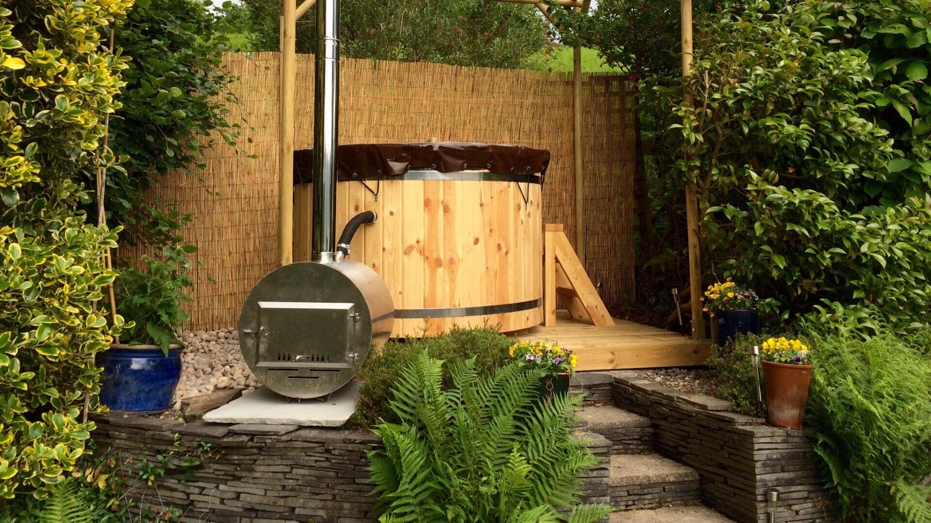 Ein hölzerner Whirlpool mit einem Holzofen aus Metall steht auf einer Holzterrasse, umgeben von üppigen Grünpflanzen, Farnen und Topfblumen, mit einem Bambusschirm und einer Steinmauer im Hintergrund.