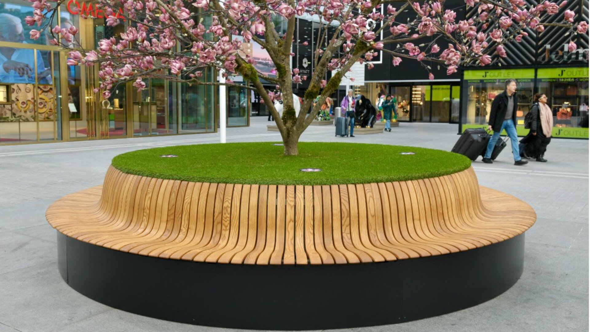 A round, modern wooden bench encircles a grassy planter with a blooming pink tree in an urban plaza; people walk by in the background.