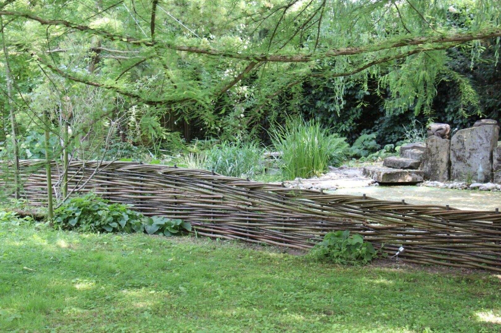 Ein rustikaler, geflochtener Holzzaun grenzt an einen üppigen Garten mit grünem Gras, blattreichen Pflanzen und hohen Bäumen. Im Hintergrund befinden sich Steine und dichtes Grün, die eine ruhige, natürliche Atmosphäre schaffen.