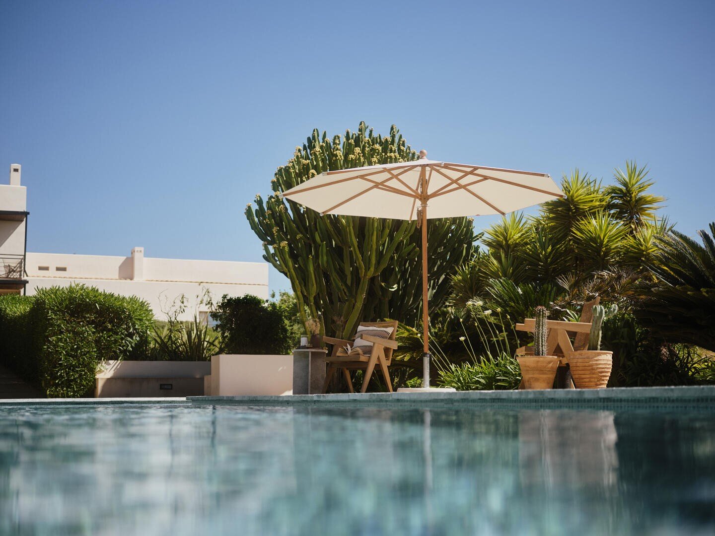 Eine Szene am Pool mit einem großen weißen Sonnenschirm, der Holzstühle und -tische beschattet, umgeben von üppigen grünen Pflanzen und Kakteen, mit einem modernen weißen Gebäude im Hintergrund unter einem klaren blauen Himmel.
