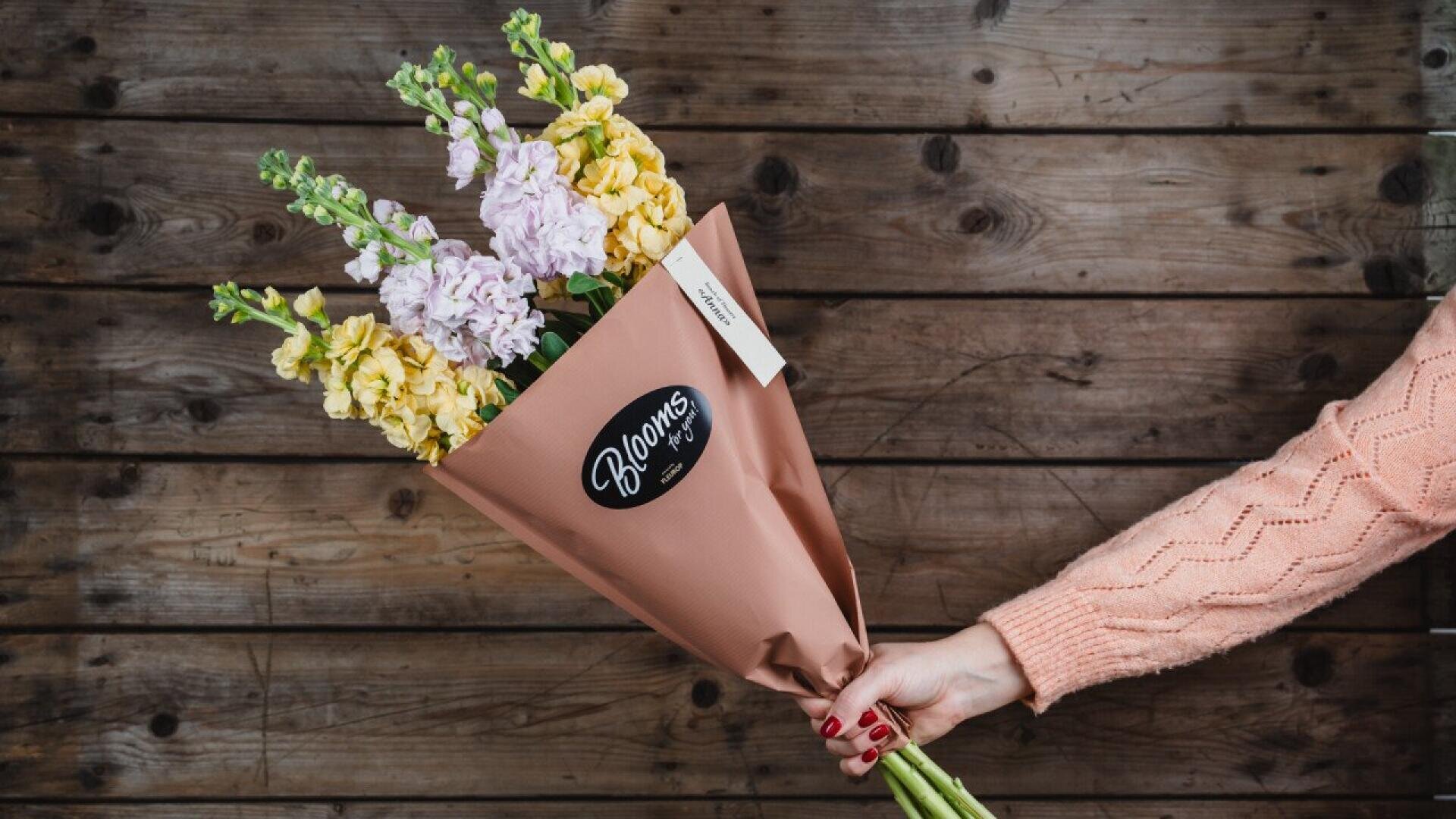 A hand in a peach sweater holds a bouquet of yellow and pale purple flowers wrapped in brown paper with a Blooms sticker, against a rustic wooden background.