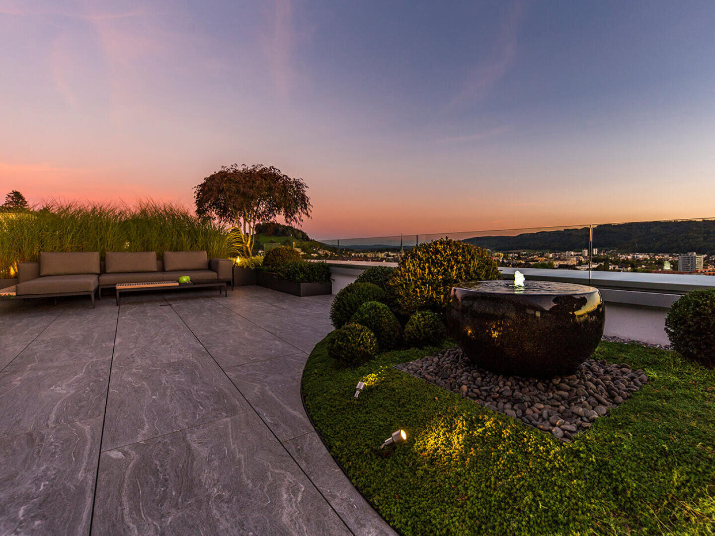 Moderne Dachterrasse bei Sonnenuntergang mit einem steinernen Wasserspiel, Landschaftssträuchern, einer Bank, hohem Gras und fernen Hügeln unter einem bunten Himmel. Der Bereich ist mit weichen Bodenleuchten beleuchtet.
