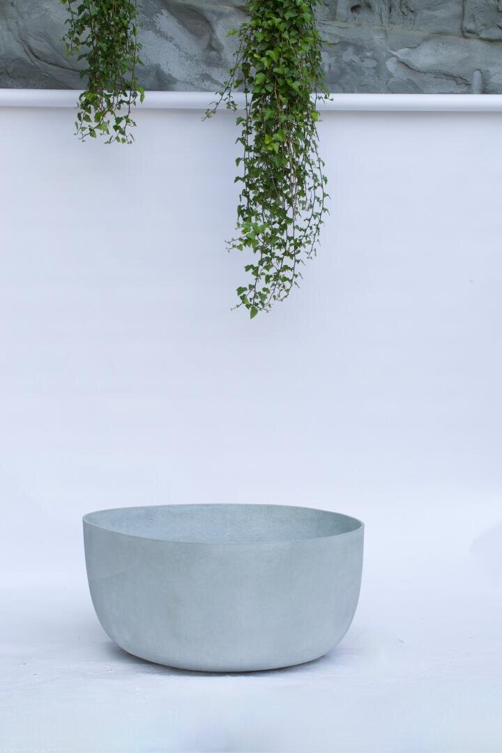 A large, light gray, round planter stands on a white surface with green tendrils hanging down from a plain, light background.
