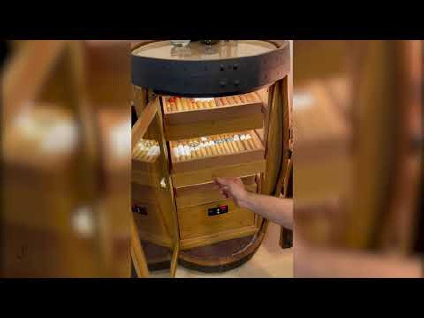 Die Hand einer Person greift in einen hölzernen Humidor mit Glastüren, in dem mehrere Reihen von Zigarren in Regalen gelagert sind.