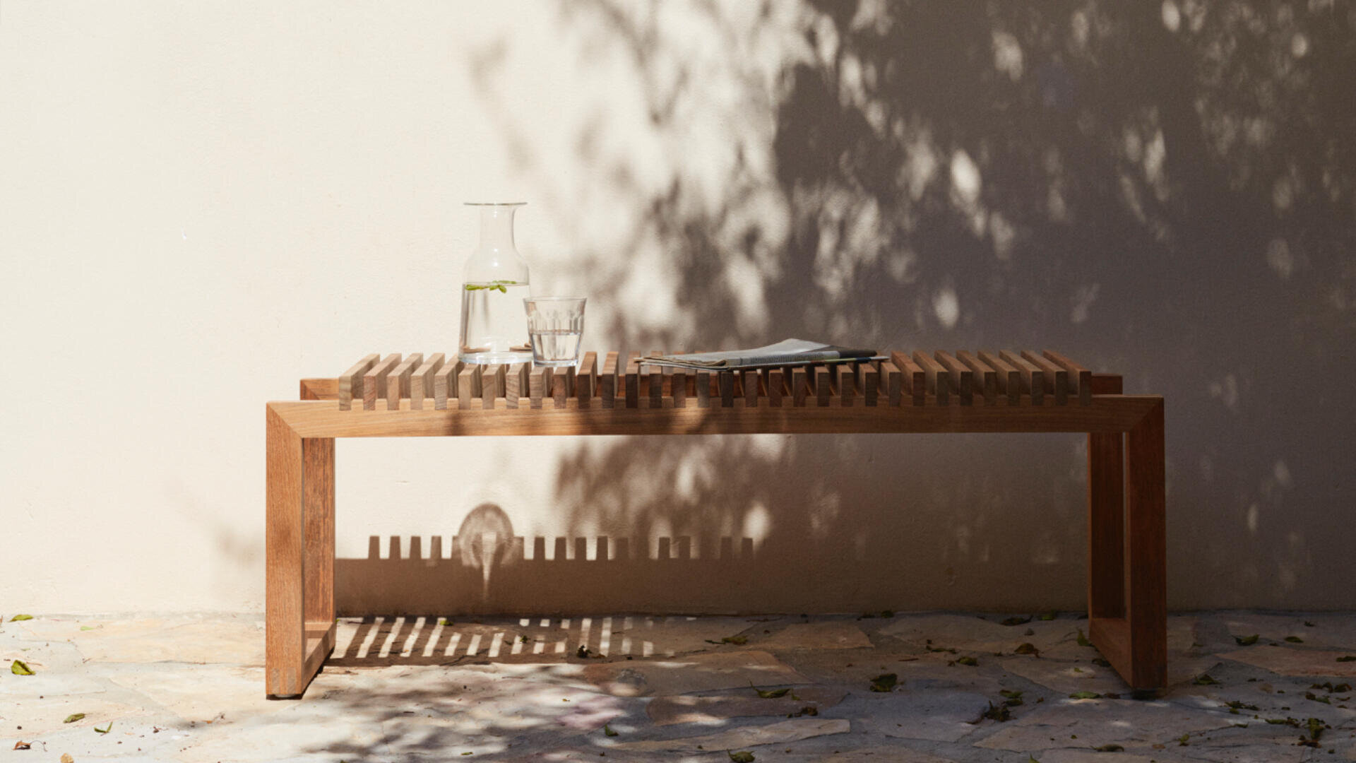Eine Holzbank mit Lattenrost steht auf einer Steinterrasse vor einer sonnenbeschienenen Wand und wirft gestreifte Schatten. Auf der Bank stehen eine Glaskaraffe mit Wasser und Minze, ein Glas Wasser und eine gefaltete Zeitung. Die Schatten der Bäume fallen auf die Szene.