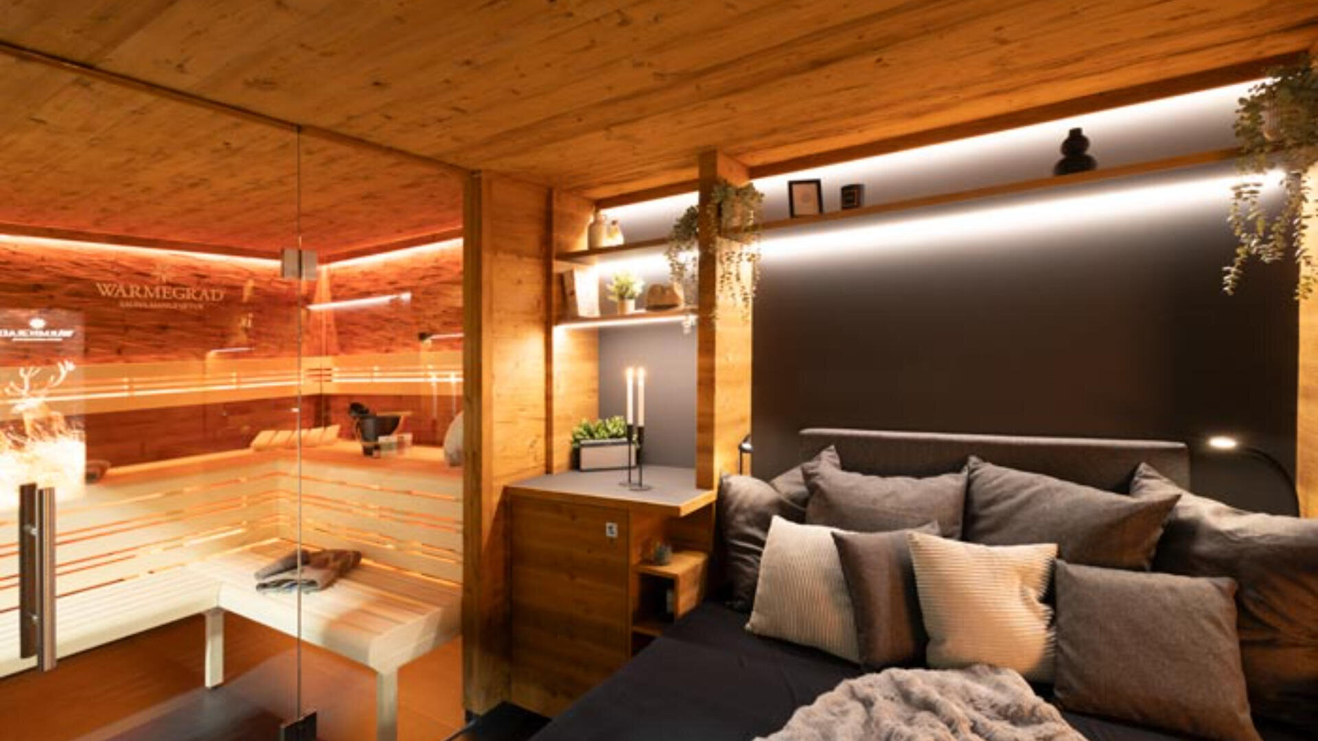Ein gemütliches, modernes Schlafzimmer mit Holzwänden, stimmungsvoller Beleuchtung, grauer Plüschbettwäsche und Regalen mit Kerzen und Pflanzen. Eine Glaswand gibt den Blick frei auf eine angrenzende Sauna mit warmer Beleuchtung und Holzbänken.
