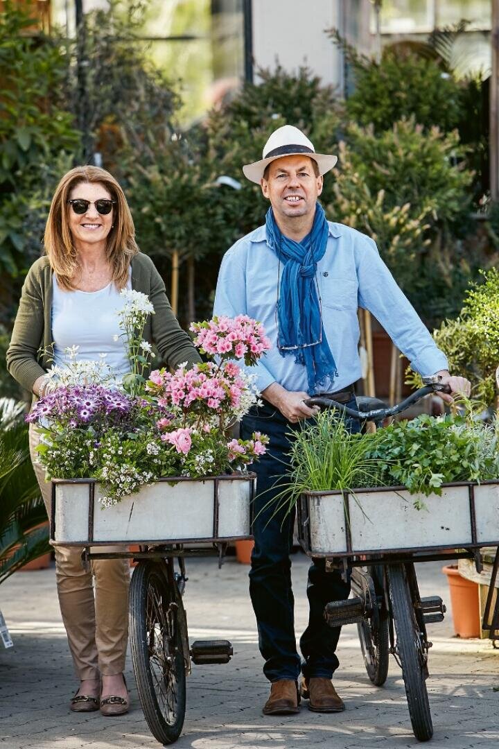 Eine lächelnde Frau und ein Mann stehen im Freien neben Fahrrädern mit Körben voller bunter Blumen und grüner Pflanzen. Der Mann trägt einen Hut und einen blauen Schal, die Frau eine Sonnenbrille und eine grüne Strickjacke. Im Hintergrund ist üppiges Grün zu sehen.