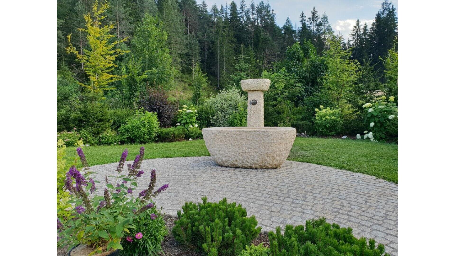 A round stone fountain stands on a cobblestone circle, surrounded by lush green plants, flowering bushes, and tall trees in a garden under a partly cloudy sky.