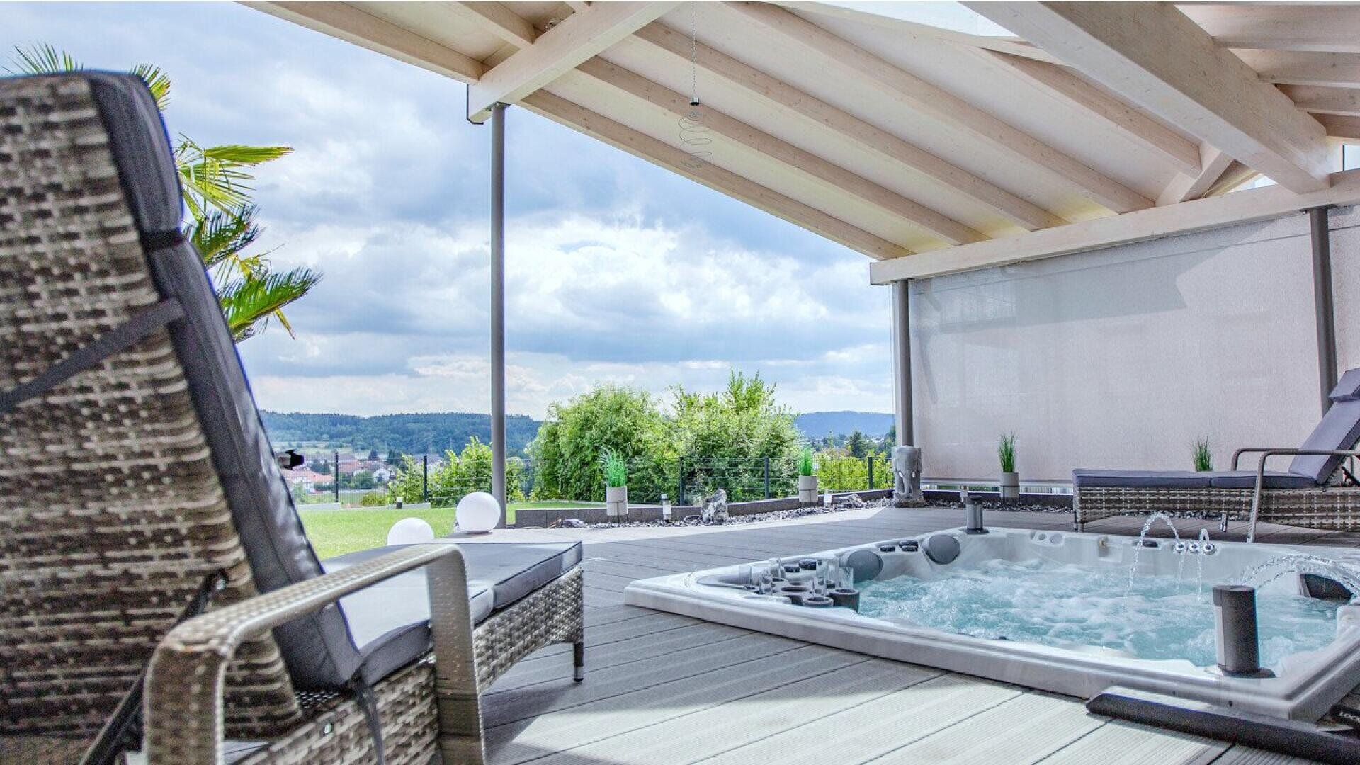 Modern outdoor patio with a hot tub bubbling, lounge chairs, potted plants, and partial wooden roof, overlooking a landscape of green hills and cloudy sky.