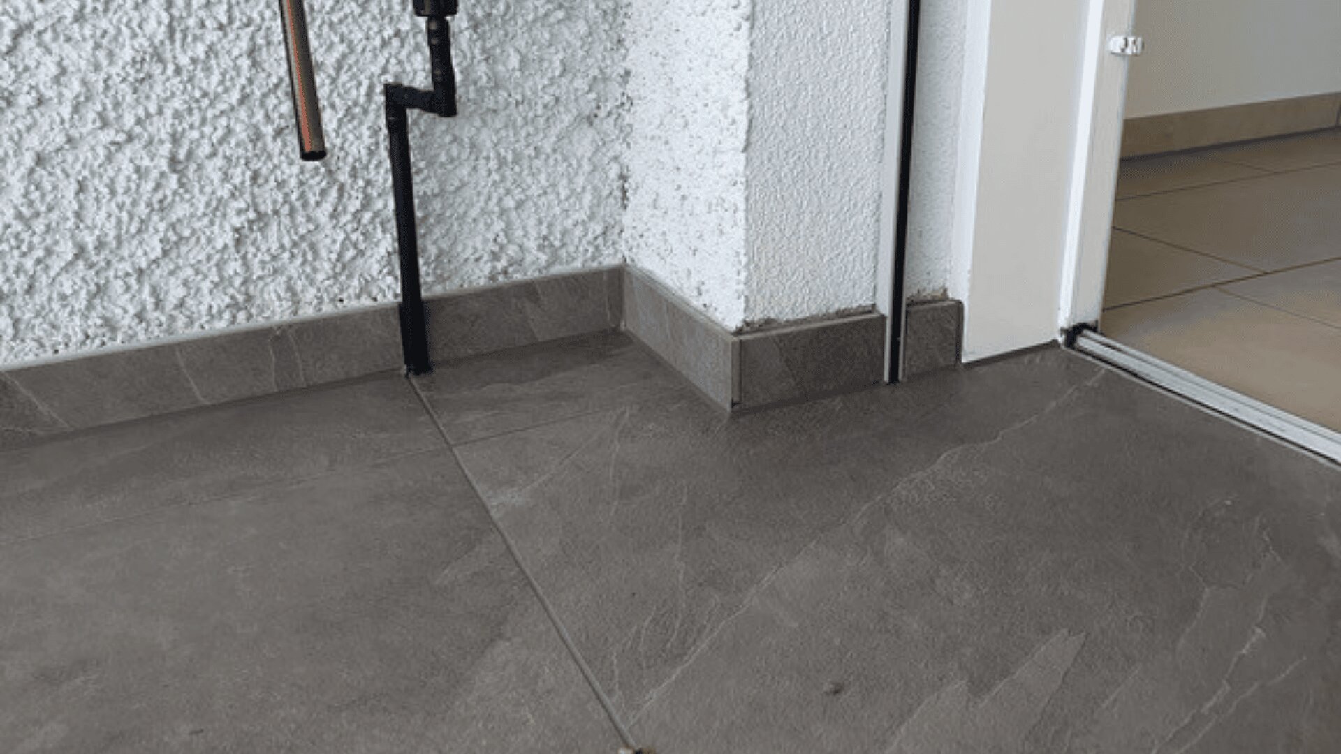Eine Zimmerecke mit strukturierten weißen Wänden, grauen Bodenfliesen, schwarzen Rohren, die an der Wand entlang verlaufen, und einer offenen Tür, die in einen Raum mit beigen Bodenfliesen führt.