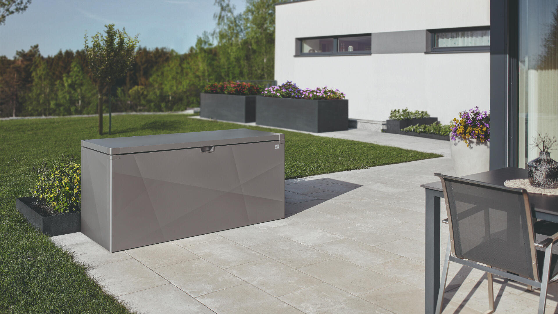 Eine schlichte, graue Aufbewahrungsbox für den Außenbereich steht auf einer Terrasse mit Topfblumen, einem Tisch und Stühlen sowie einem modernen weißen Haus, umgeben von grünem Gras und Bäumen im Hintergrund.