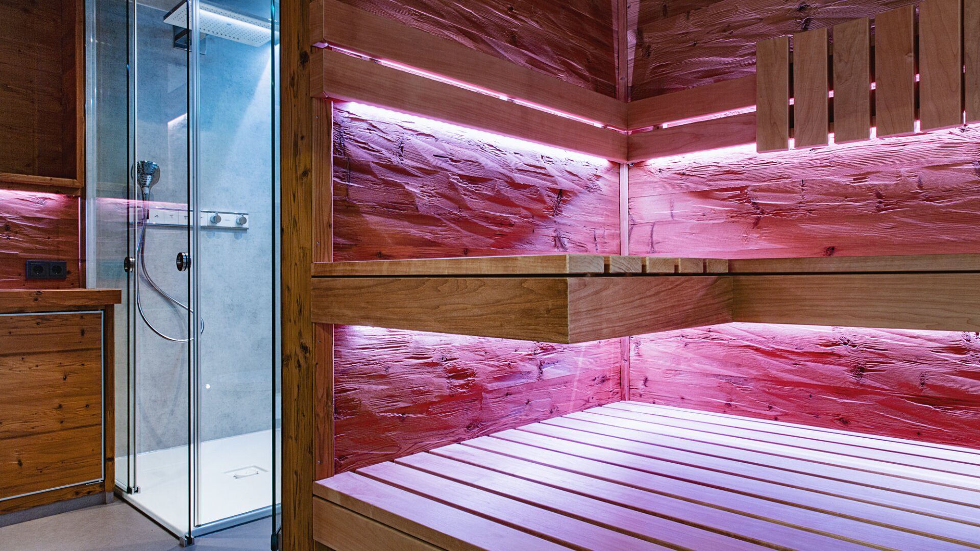 Moderne Holzsauna mit rosafarbener LED-Beleuchtung, mit zweistufigen Bänken, strukturierten Holzwänden und einem glasüberdachten Duschbereich im Hintergrund. Der Raum wirkt sauber und zeitgemäß.