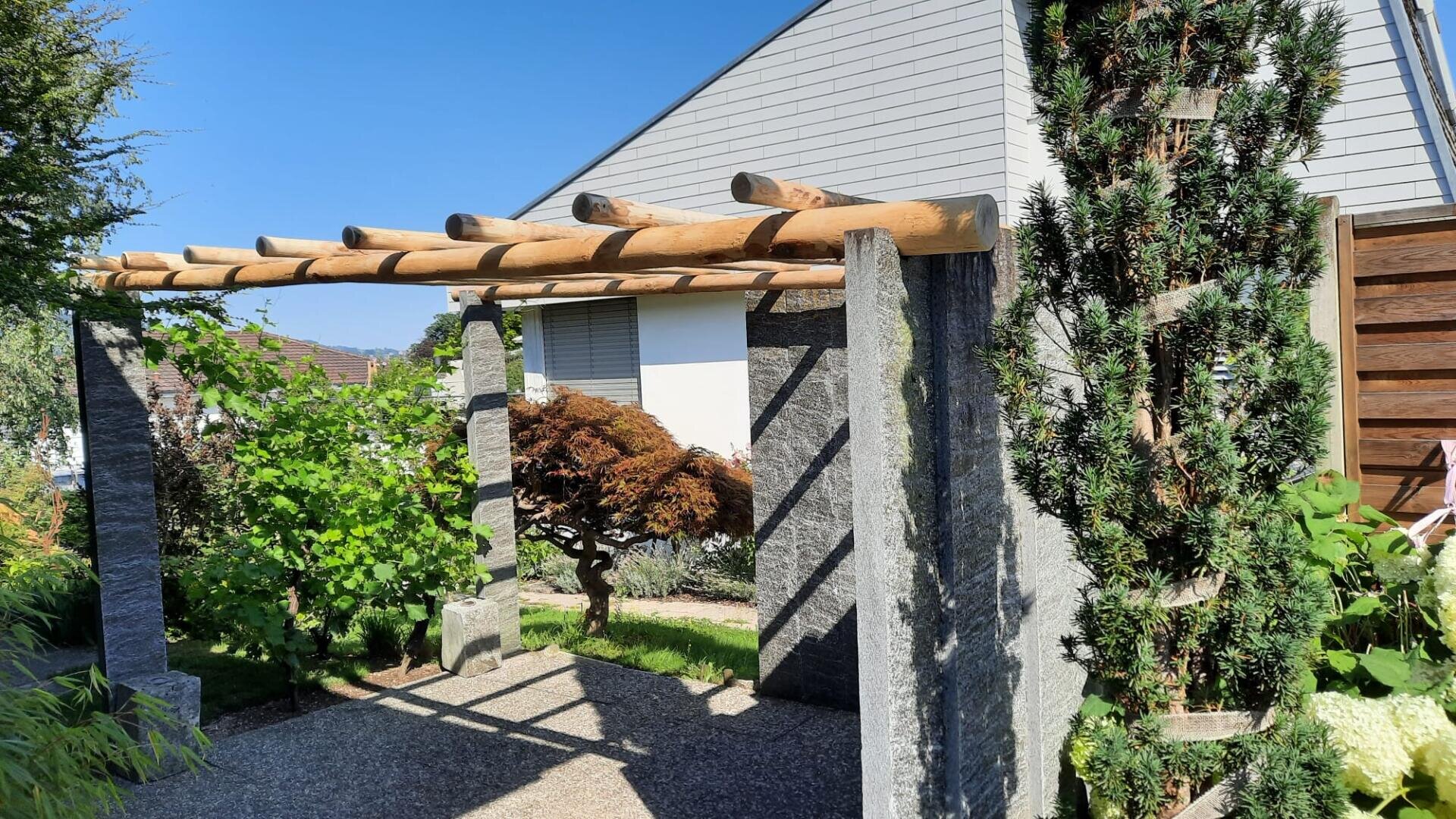 Eine hölzerne Pergola mit Steinsäulen steht in einem sonnenbeschienenen Garten, umgeben von grünen Pflanzen und Bäumen, neben einem modernen Gebäude mit grauer Fassade unter einem klaren blauen Himmel.