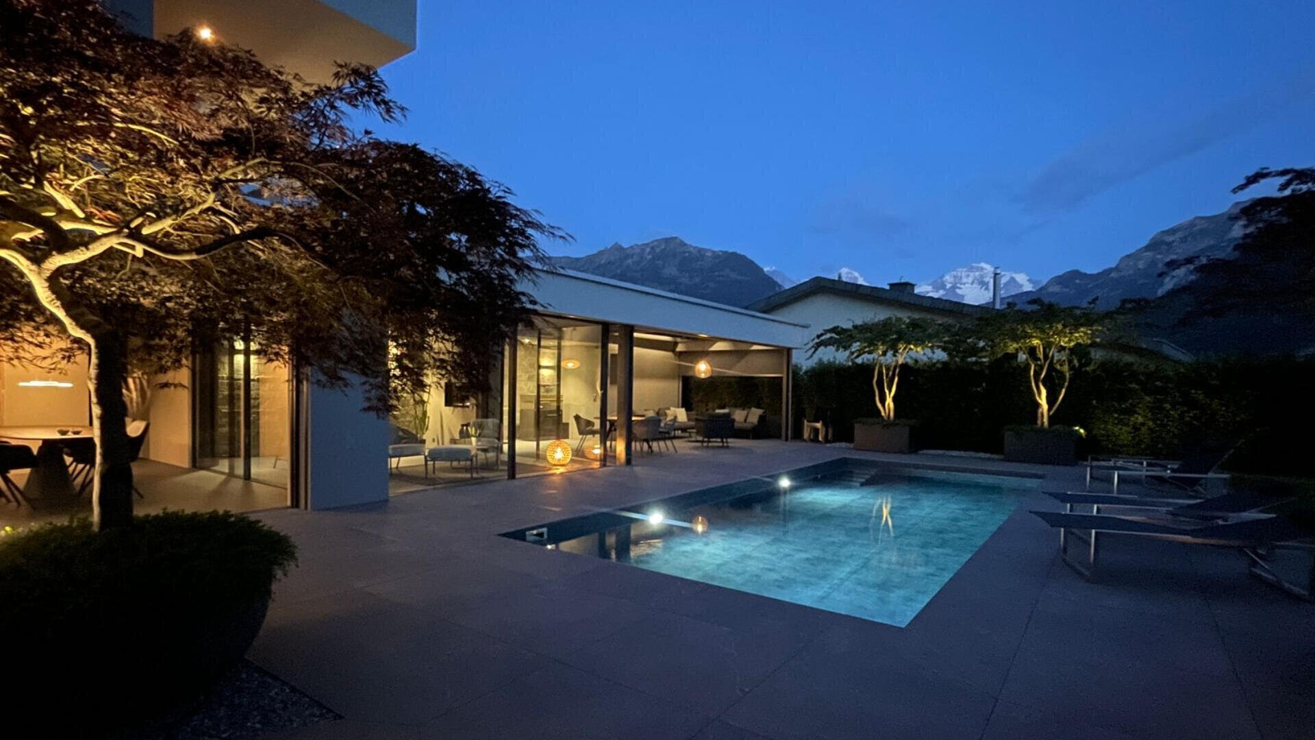 Modernes Haus mit großen Fenstern und einem beleuchteten Swimmingpool im Hinterhof in der Abenddämmerung. Terrassenstühle säumen den Pool, und im Hintergrund sind die Berge unter dem Dämmerungshimmel zu sehen. Bäume und Umgebungsbeleuchtung schaffen eine gemütliche Atmosphäre.