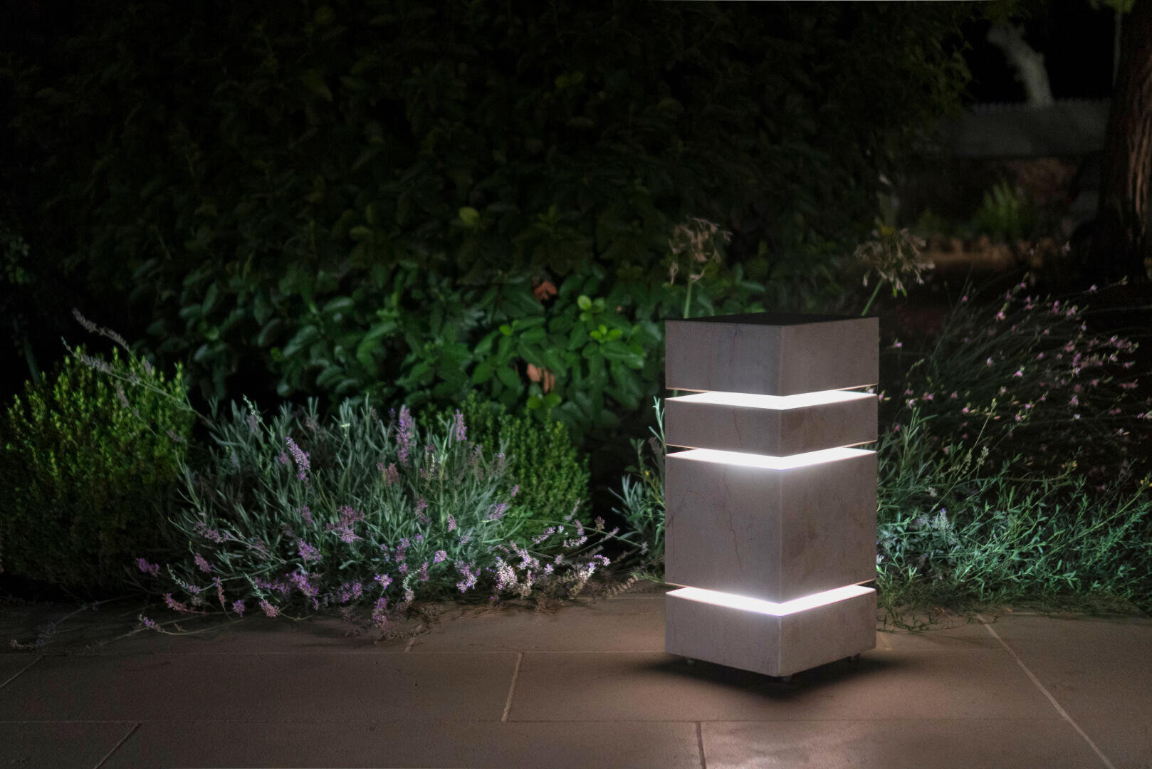 Eine moderne, rechteckige Gartenleuchte leuchtet in der Nacht und beleuchtet grüne Sträucher, Lavendel und einen Steinweg in der Nähe.