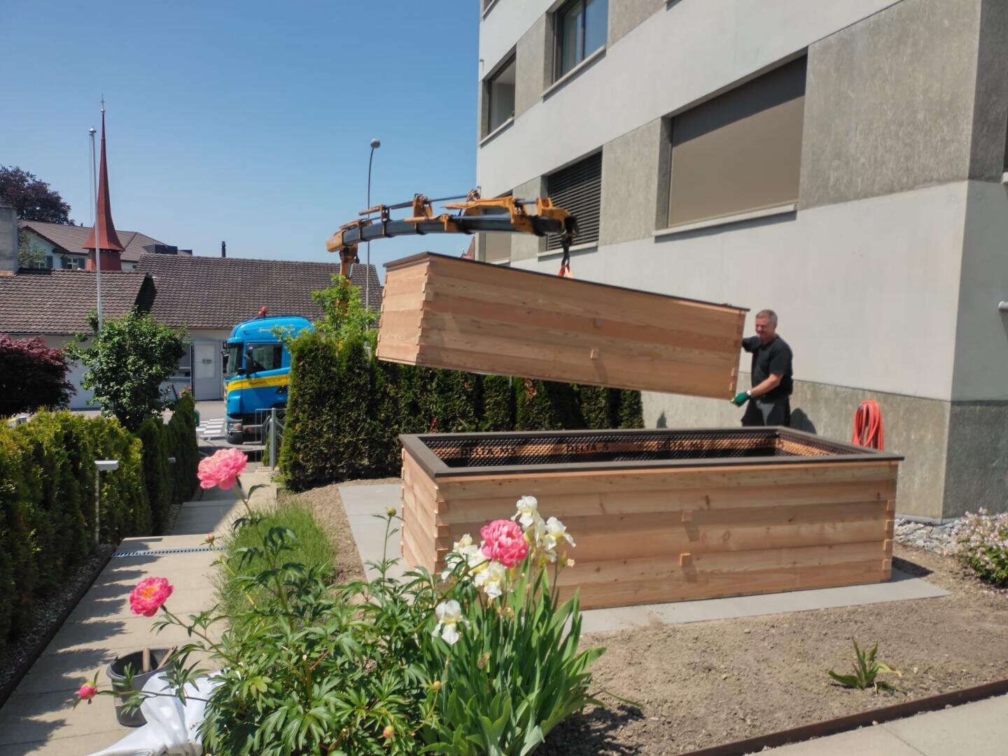 Eine Person hebt mit einem Kran den Holzdeckel von einem großen Hochbeet in der Nähe eines Gebäudes. In der Nähe blühen rosafarbene Blumen, und im Hintergrund ist an einem sonnigen Tag ein blauer Lastwagen geparkt.