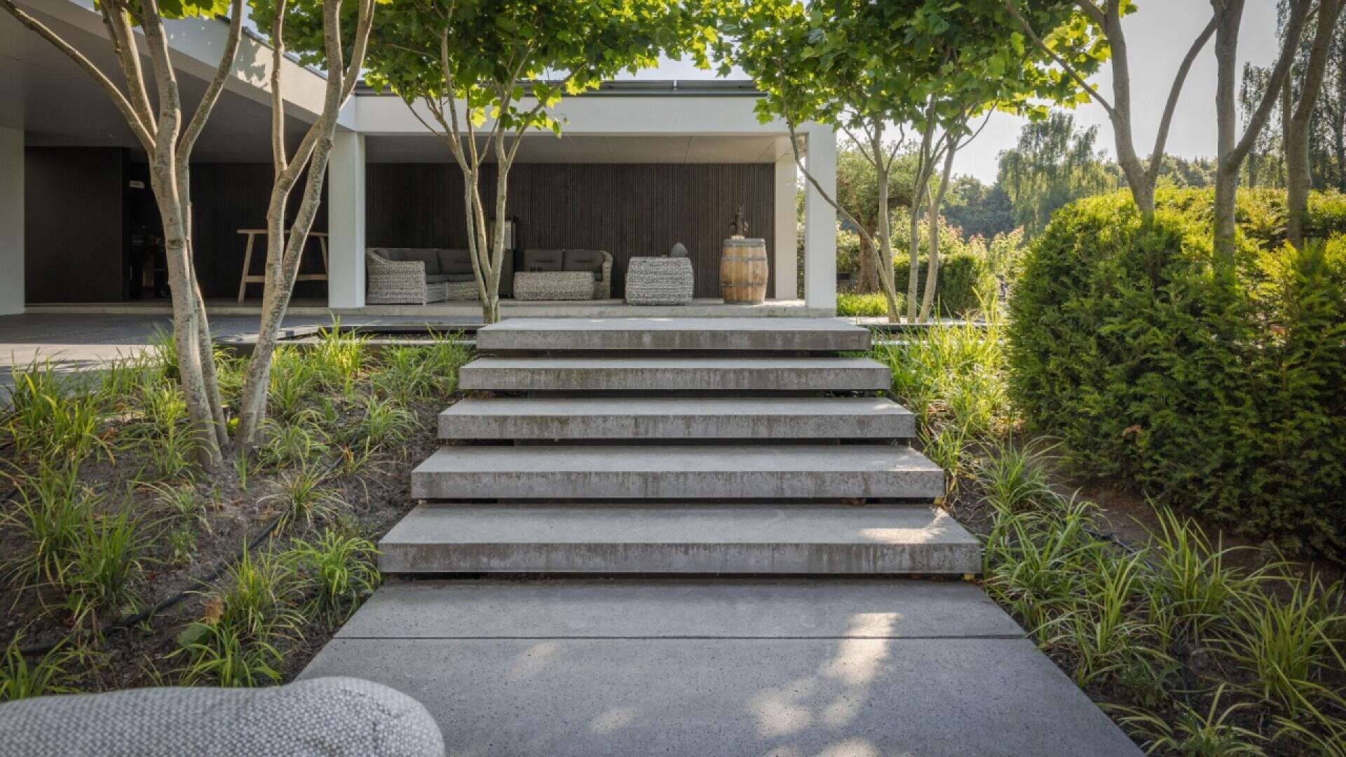 Wide concrete steps lead up through a modern landscaped garden with trees and greenery, towards a shaded patio area with outdoor seating and a wooden barrel. The scene appears peaceful and inviting.
