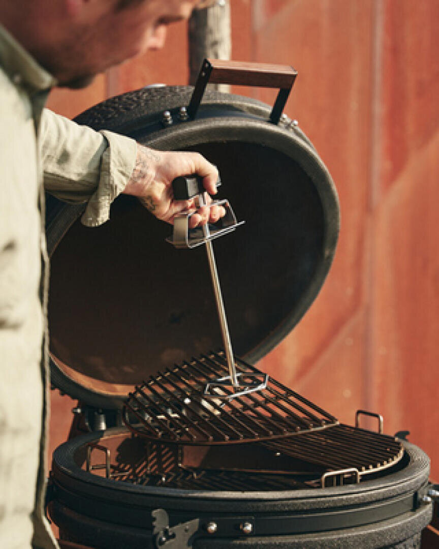 Eine Person hebt mit einem Metallwerkzeug den Grillrost eines runden, schwarzen Outdoor-Grillgeräts mit geöffnetem Deckel vor einem unscharfen braunen Hintergrund an.