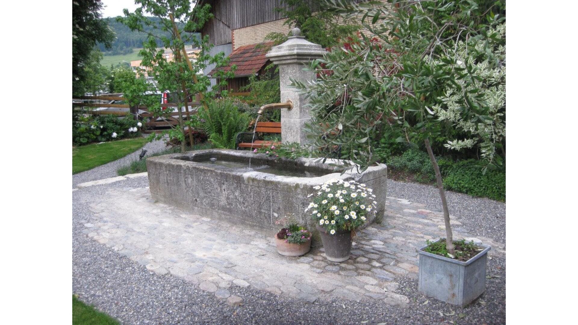 A rectangular stone fountain with a brass spout stands on a cobblestone area surrounded by plants, trees, and potted flowers in a garden setting, with benches and a wooden building in the background.