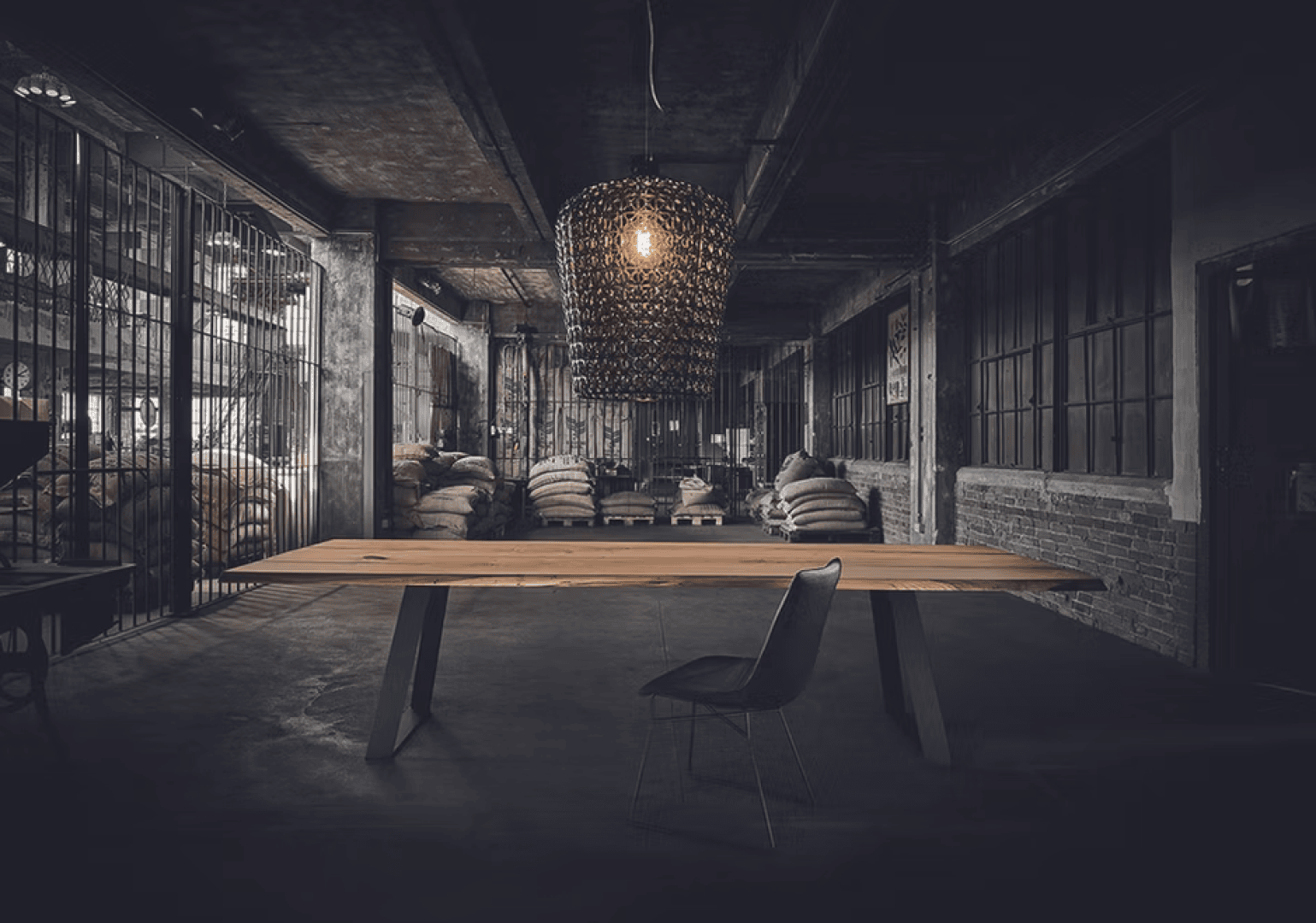 Ein einzelner Stuhl sitzt an einem langen Holztisch in einem schwach beleuchteten Industrielager mit freiliegenden Ziegelsteinen, Metallkäfigen und gestapelten Jutesäcken im Hintergrund. Eine große, moderne Pendelleuchte hängt über dem Tisch.