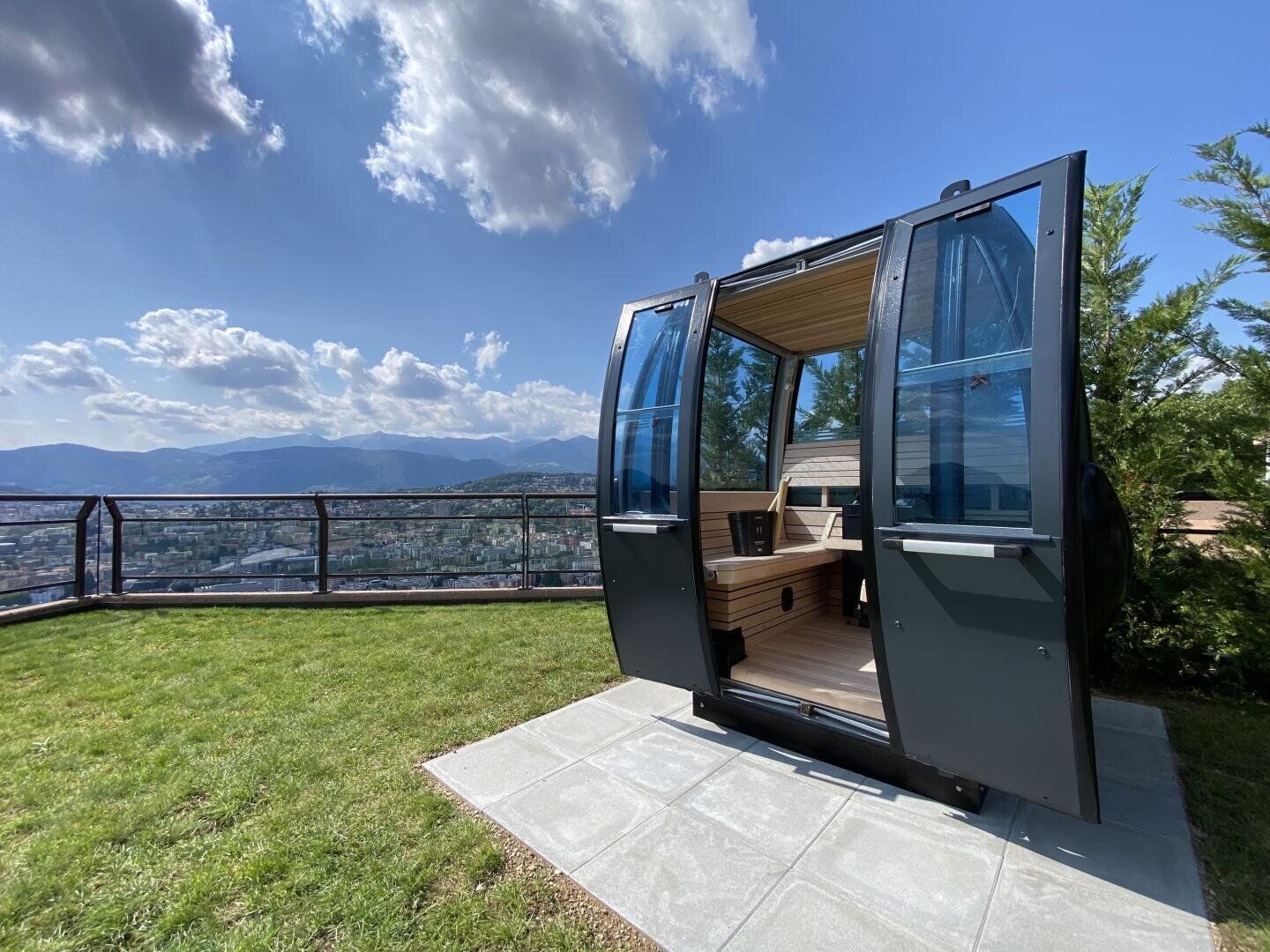 A modern, black outdoor sauna shaped like a cable car sits on a stone patio with an open door, overlooking a grassy yard, scenic mountains, and a city below under a blue sky with scattered clouds.