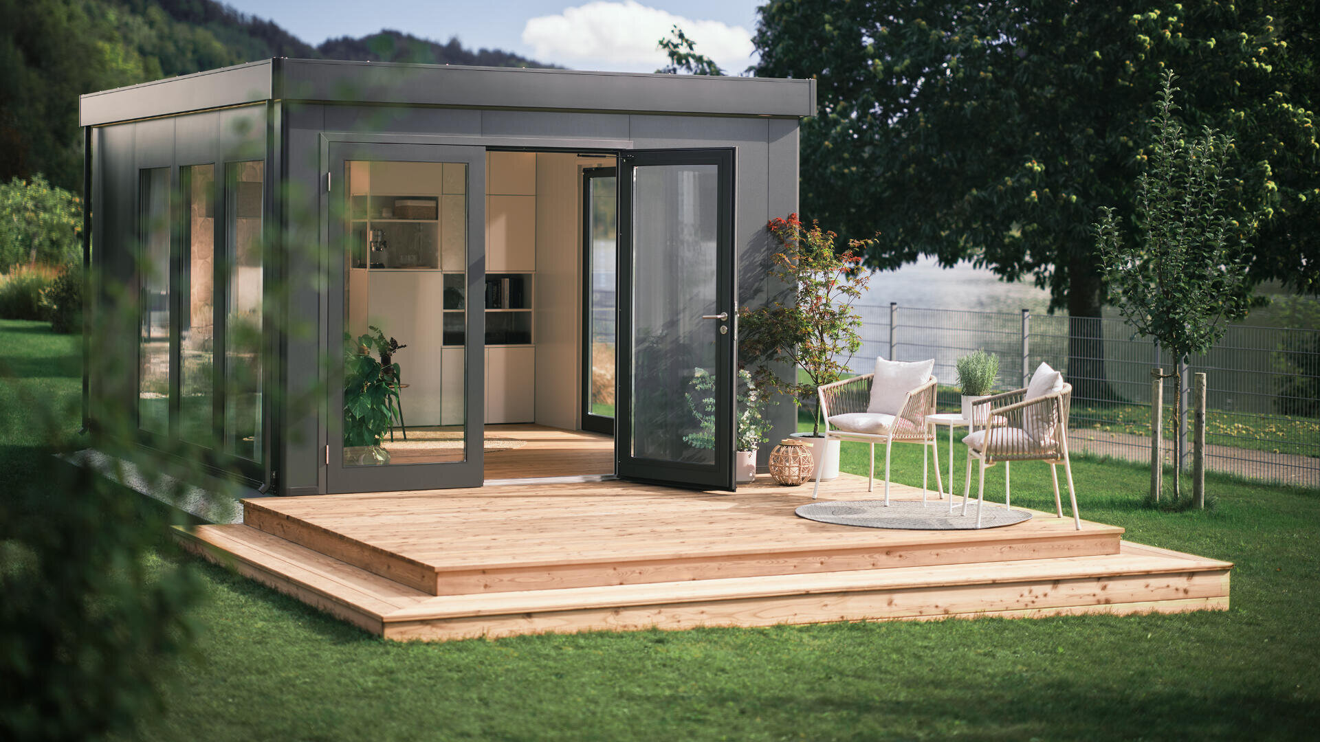 Ein modernes Gartenstudio mit großen Glastüren öffnet sich auf eine Holzterrasse mit zwei Stühlen und einem kleinen Tisch. Das Studio ist von grünem Gras, Bäumen und Pflanzen umgeben, und im Hintergrund sind Hügel zu sehen.