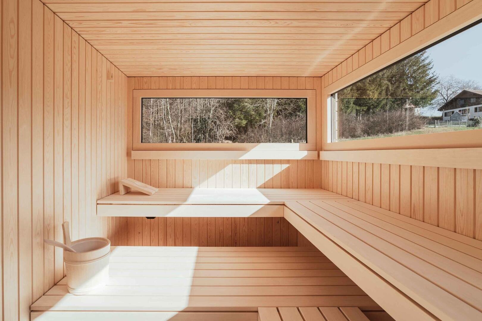 A modern wooden sauna with large windows, built-in benches, a wooden bucket with ladle and a tranquil view of trees and a house. The sunlight streams through the windows and creates a warm and inviting atmosphere.