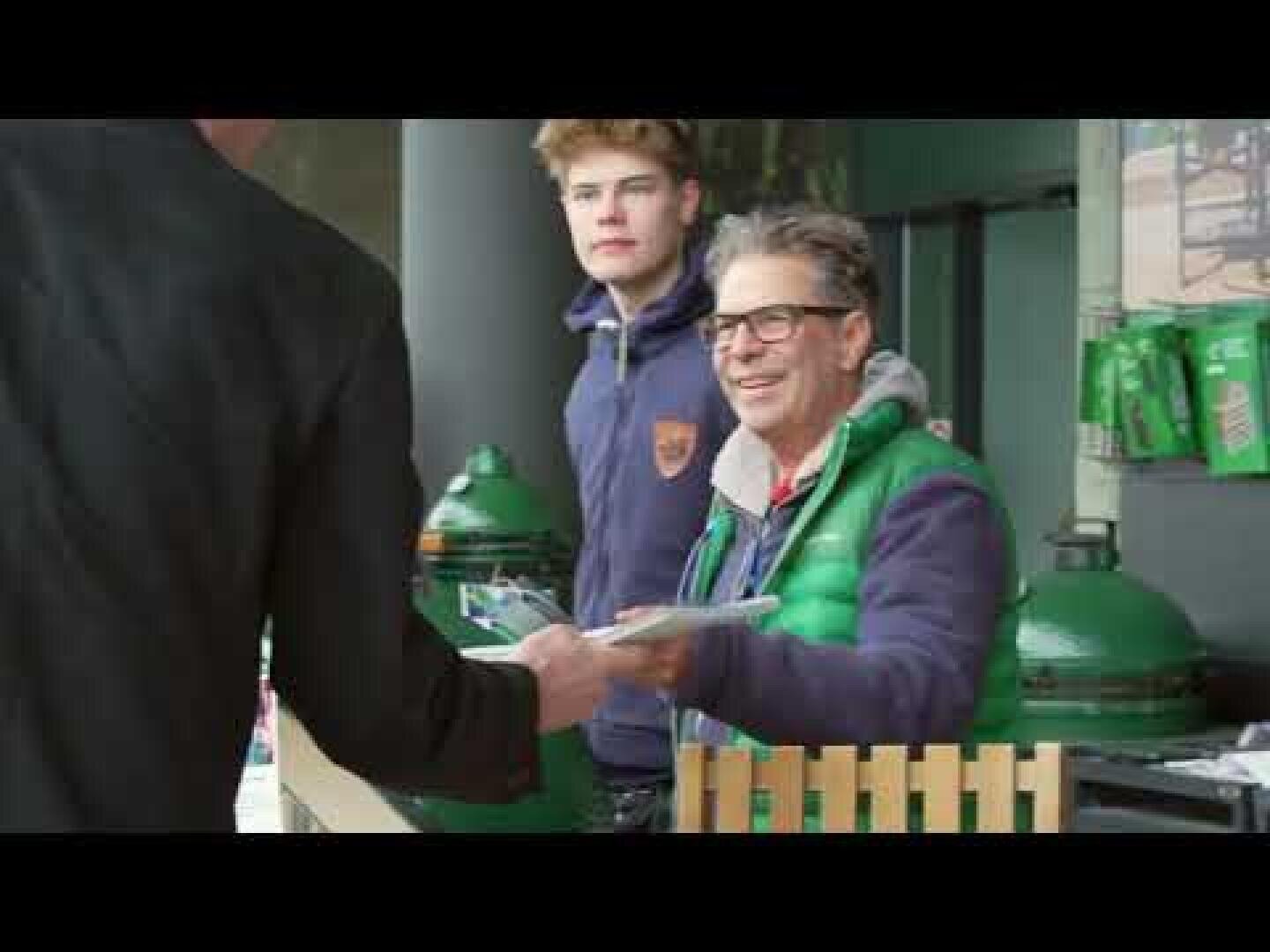 Two people are standing at a stand, one younger and one older with glasses and a green vest, while the older person hands someone outside the picture a leaflet or newspaper. Green barbecues and bags can be seen in the background.