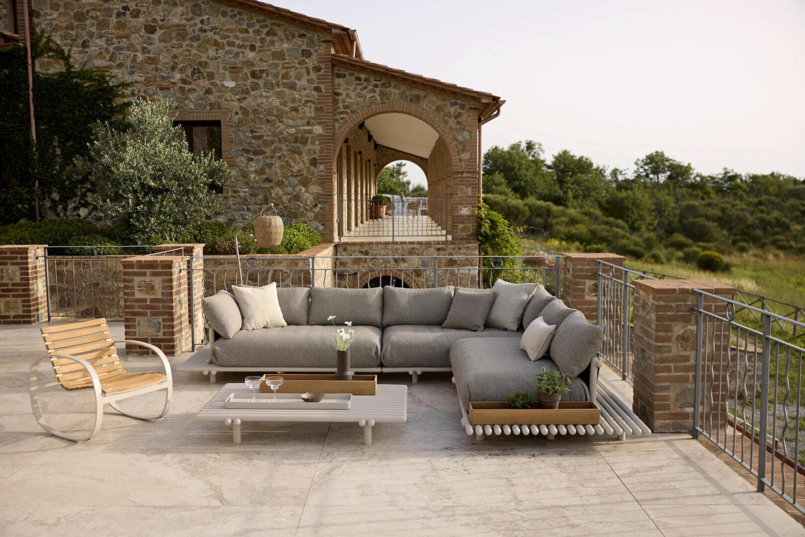 The spacious terrace with stone and brick accents features a modern sectional sofa with gray cushions, a white coffee table and a wooden lounge chair with a view of green trees and a rustic stone house.