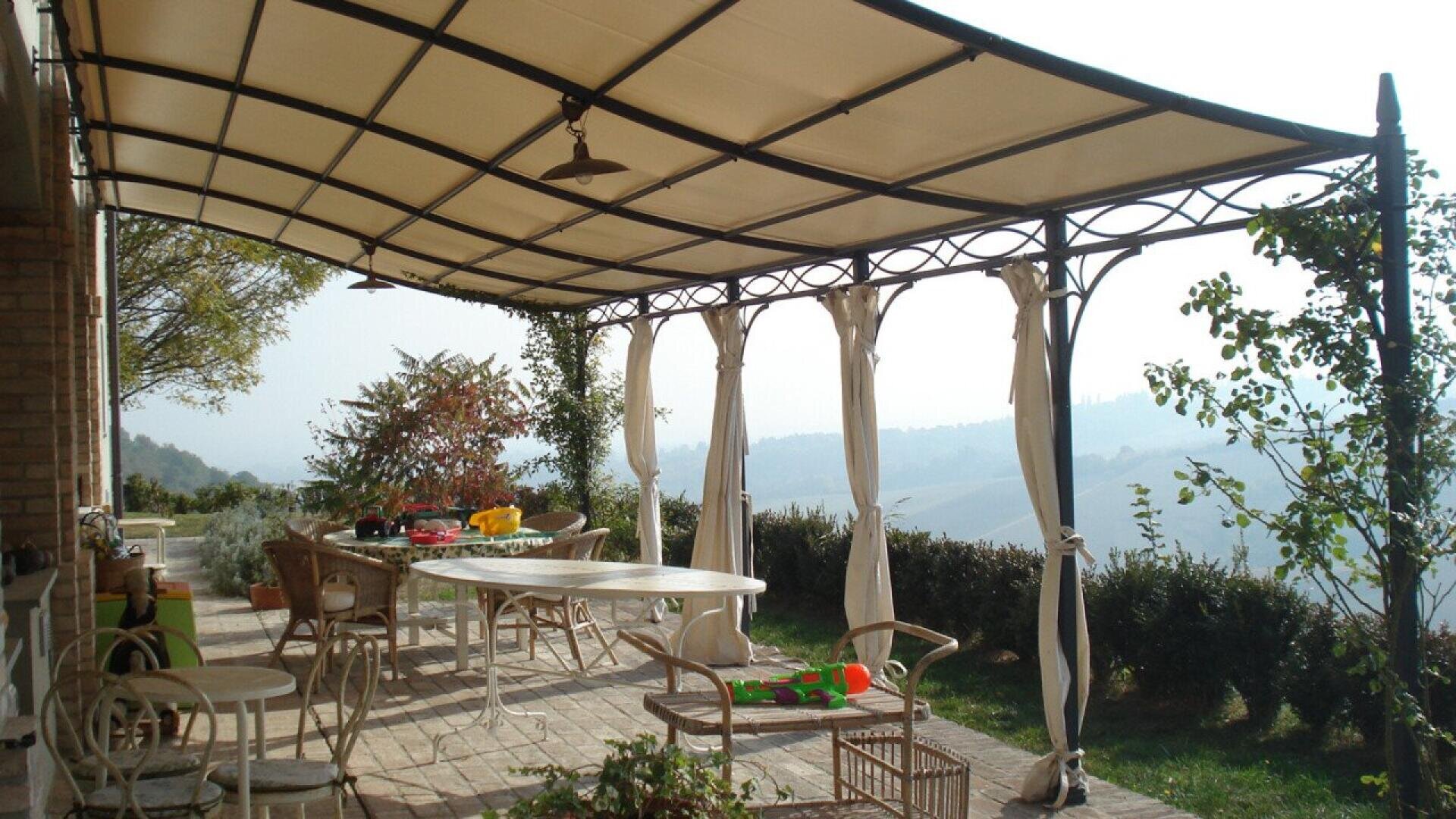 A covered patio with a beige canopy, white curtains, and outdoor furniture including a table and chairs. Childrens toys are on the floor and table. Greenery and distant hills are visible in the background.