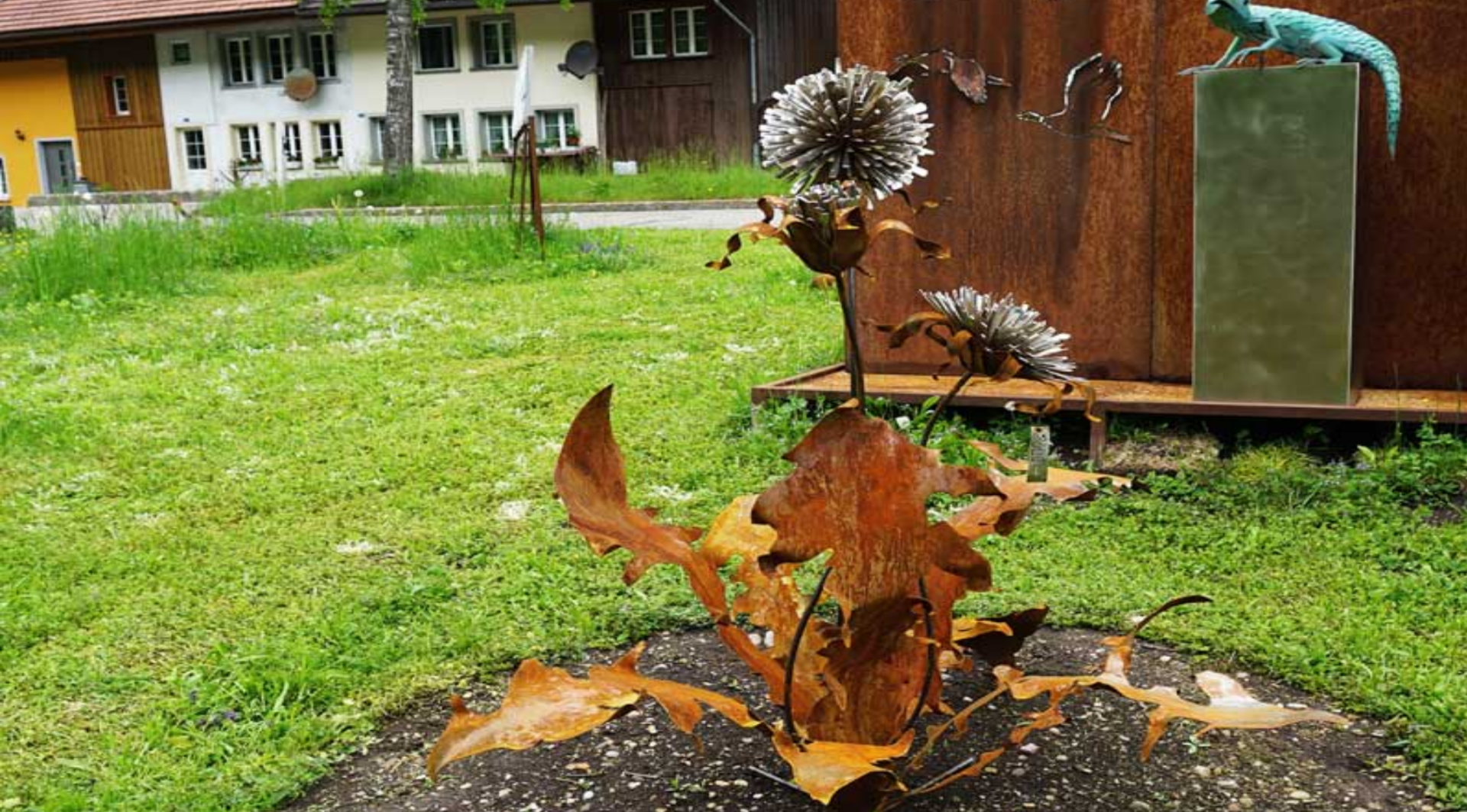 Eine rostfarbene Metallskulptur eines Löwenzahns mit großen Blättern und runden Blütenköpfen steht im Gras.
