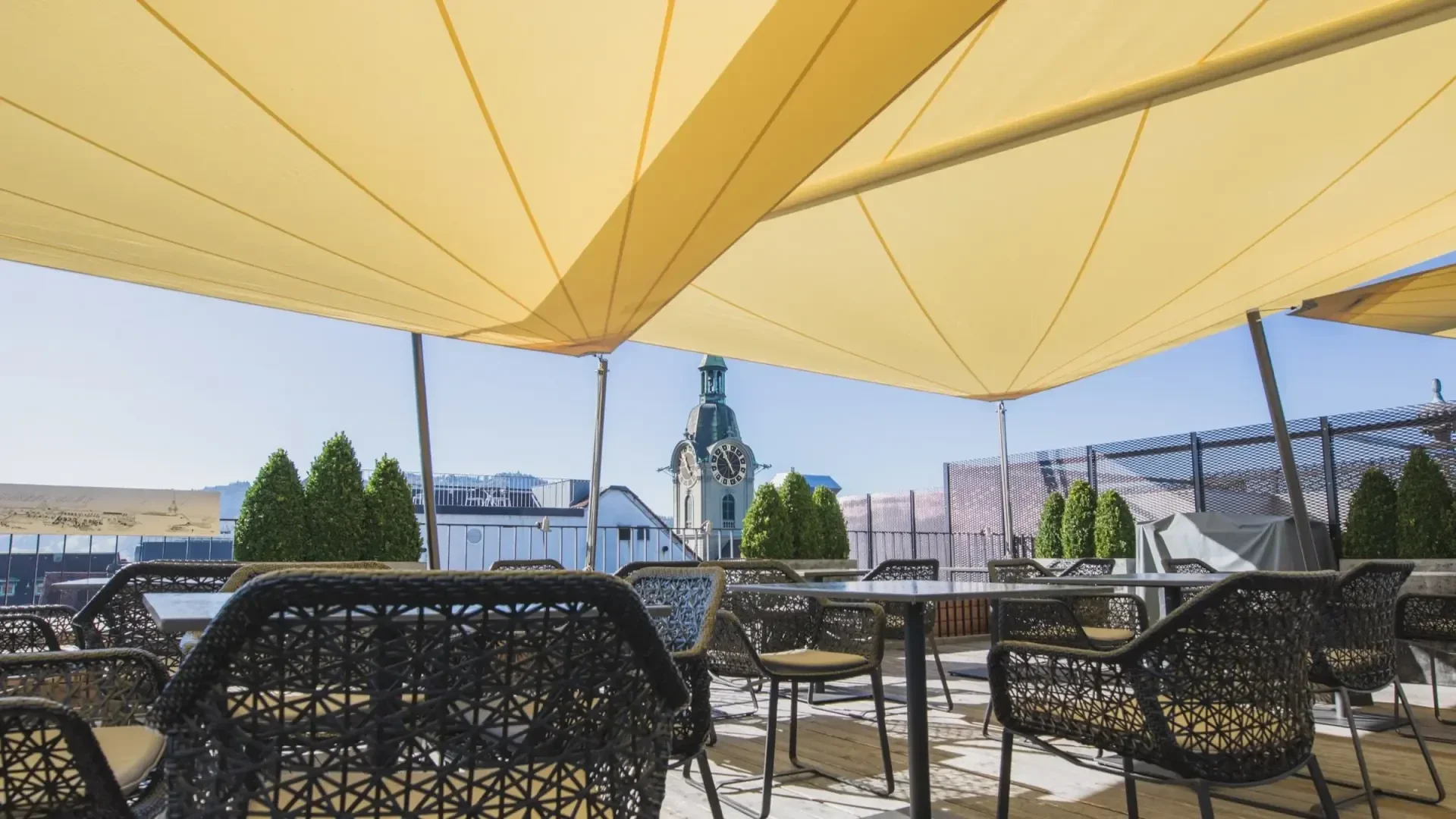 Terrace with woven chairs and tables under large yellow tarpaulins, with a view of the roofs of the city and a clock tower in the background, with a clear blue sky and a few green bushes.
