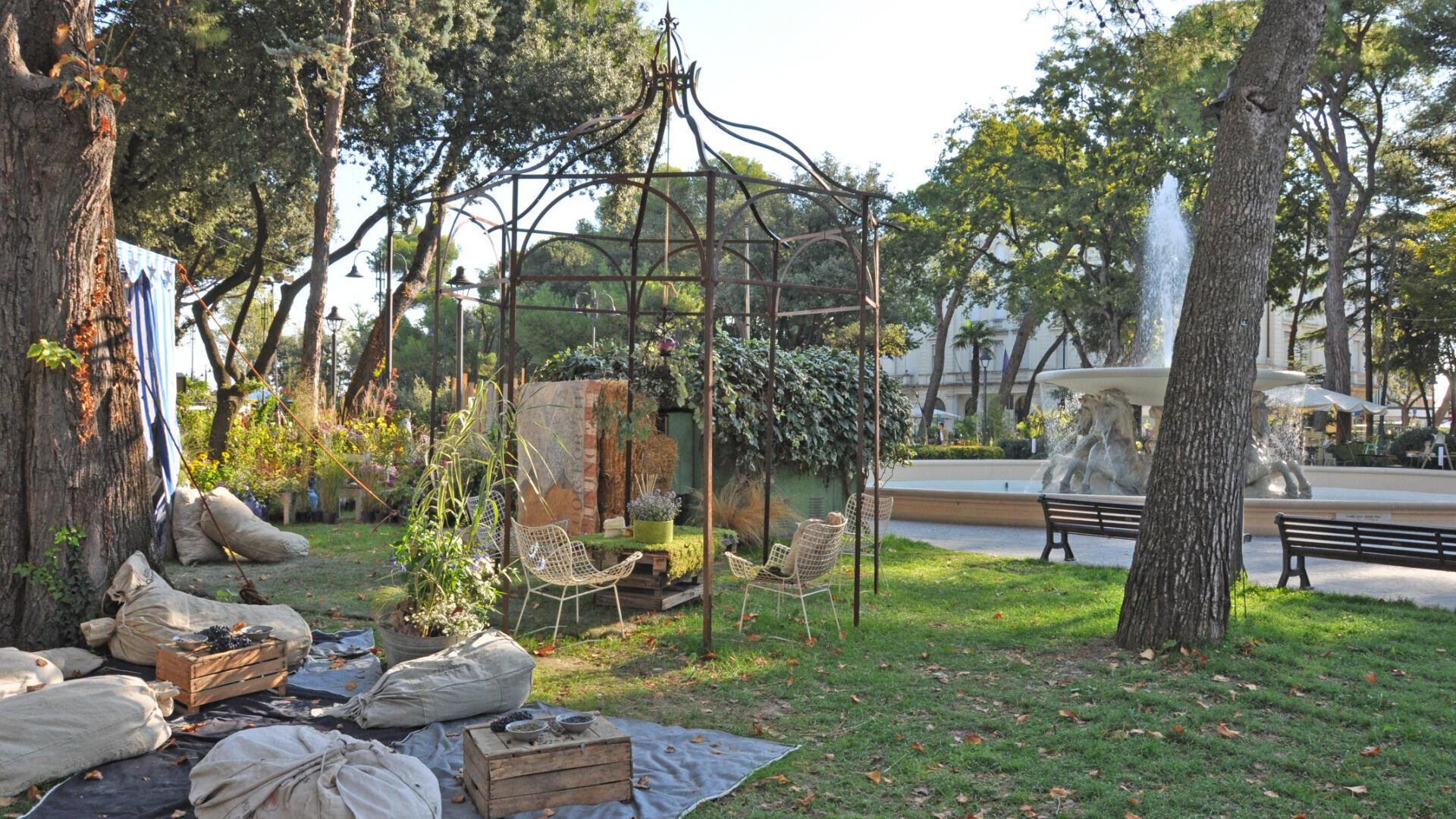 In einem Park ist ein Metallpavillon mit Stühlen und Pflanzen aufgestellt. Sitzsäcke und Holzkisten liegen in der Nähe auf dem Rasen, und im Hintergrund ist zwischen Bäumen und Bänken ein großer Springbrunnen mit Skulpturen zu sehen.