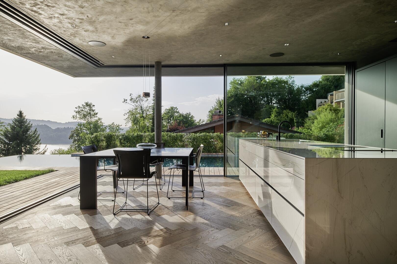 Modern open plan kitchen and dining area with large glass walls overlooking a pool, lush greenery and a lake. Marble countertops and wood floors create a stylish, modern space.