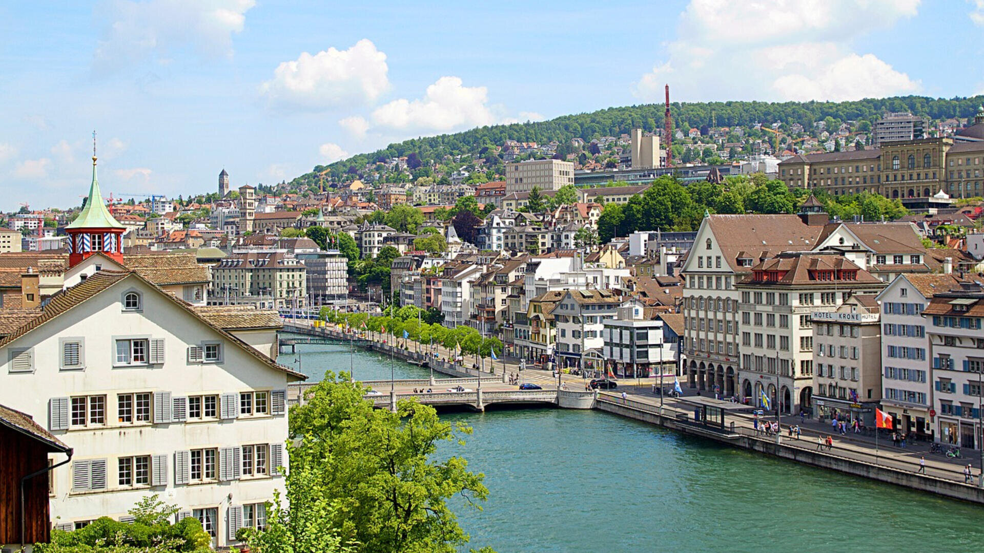 Ansicht von Zürich, Schweiz, mit historischen Gebäuden entlang des Flusses Limmat, Brücken und üppig grünen Hügeln im Hintergrund bei teilweise bewölktem Himmel.