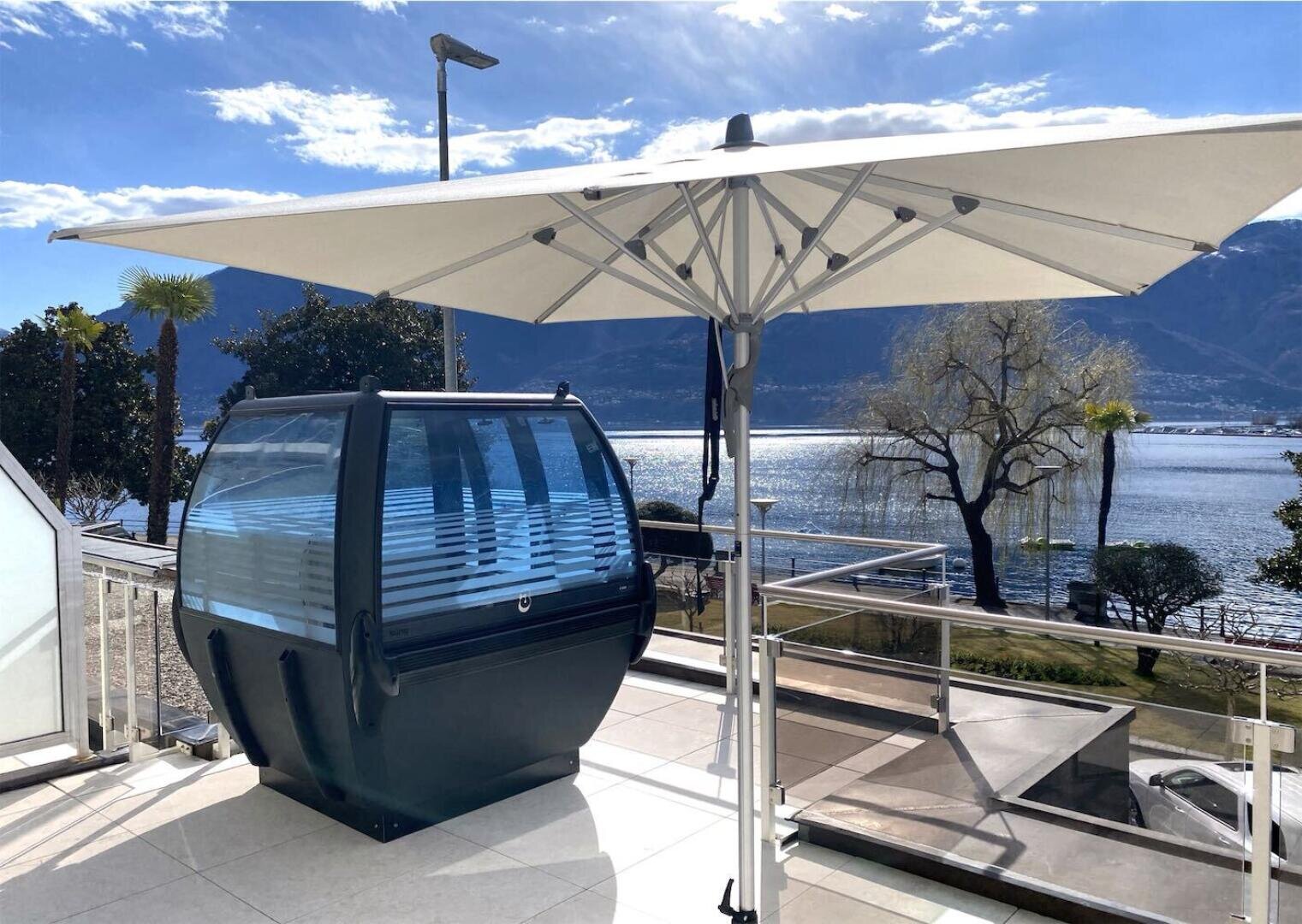 A large white patio umbrella shades a unique glass and metal cabin-like pod on a sunny terrace overlooking a scenic lake and mountains in the background.