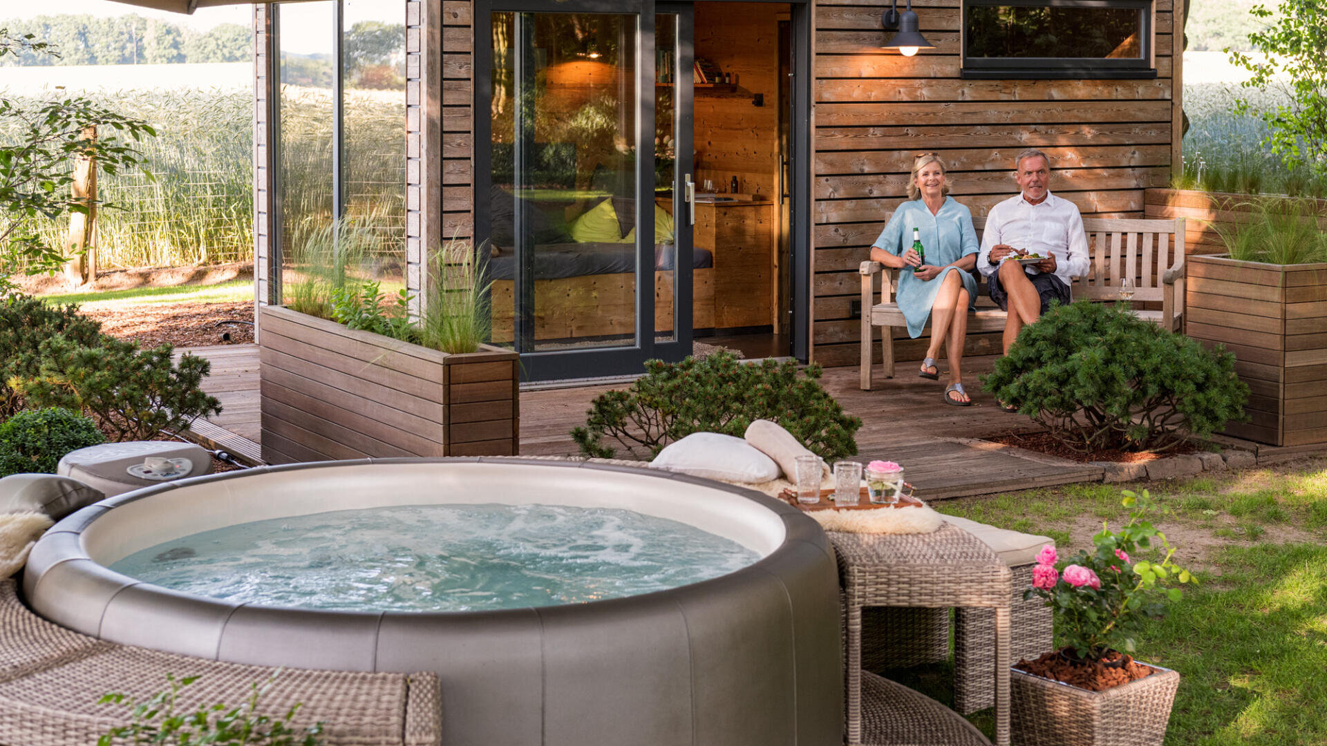 Ein älteres Ehepaar sitzt auf einer Holzbank auf seiner Terrasse, lächelt und genießt ein Getränk. Im Vordergrund befindet sich ein sprudelnder Whirlpool mit einem kleinen Tisch mit Erfrischungen und Blumen in der Nähe. Die Szene ist gemütlich und von viel Grün umgeben.